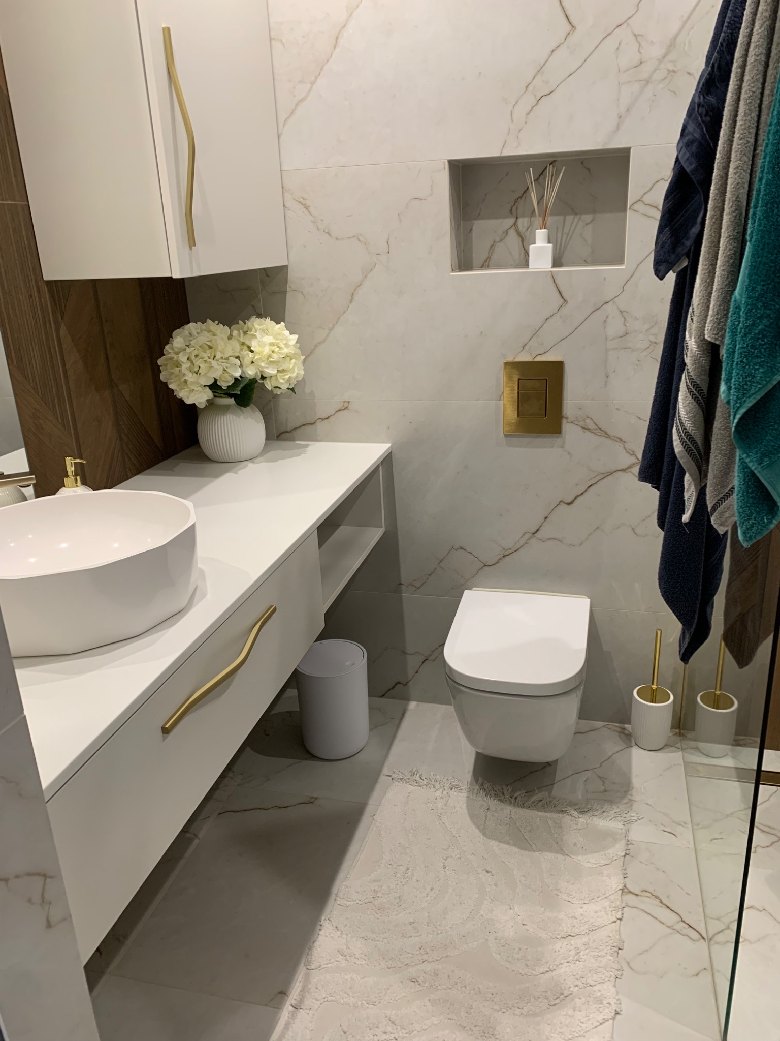 Nowoczesna, jasna łazienka po remoncie w Jasionce, z marmurowymi płytkami, wiszącą toaletą, białą umywalką nablatową i złotymi akcentami.