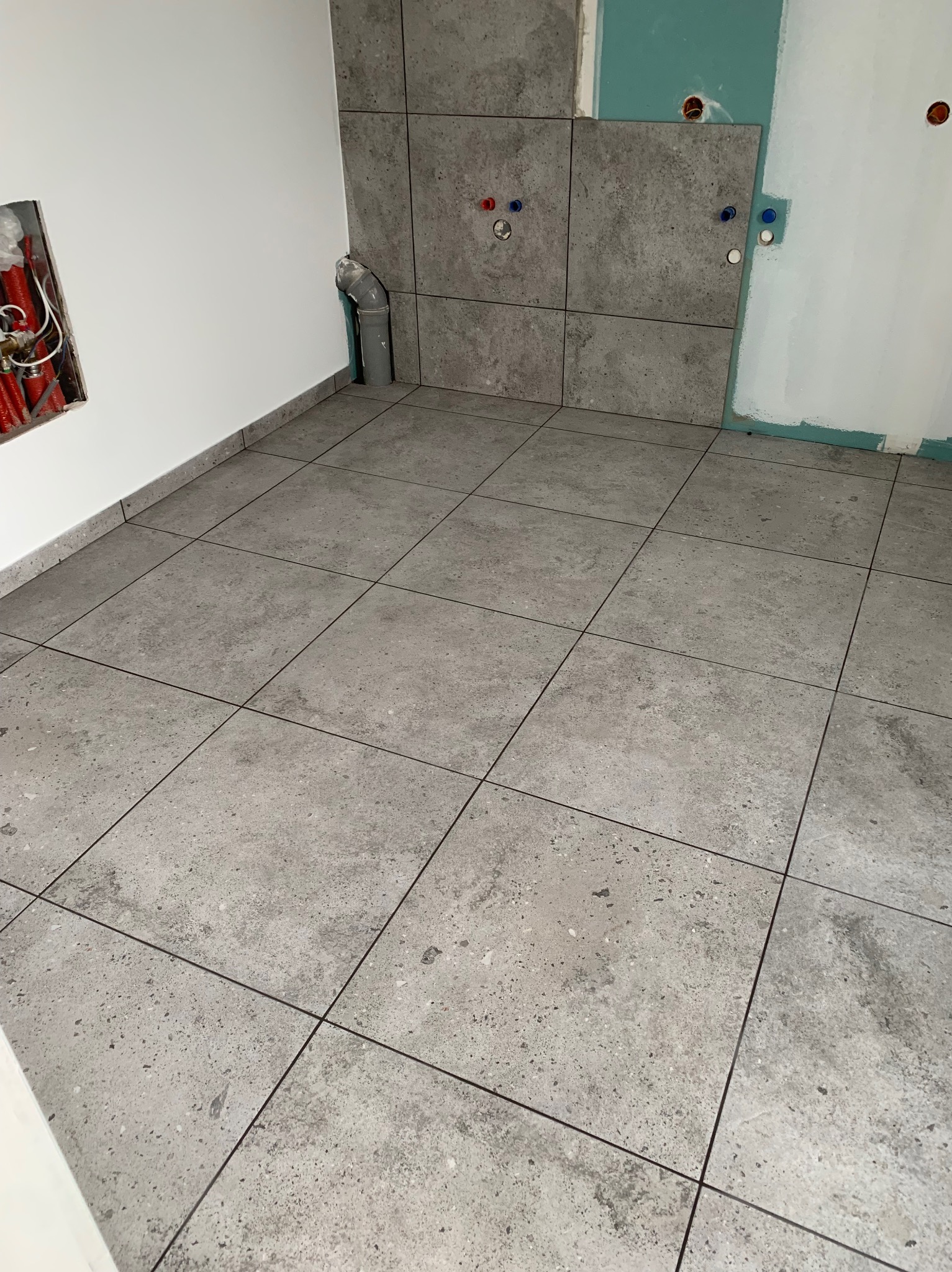 Łazienka w trakcie remontu: ułożone szare płytki podłogowe i ścienne, widoczne instalacje wodne i elektryczne w ścianach, ściana z płyt kartonowo-gipsowych w trakcie wykańczania.