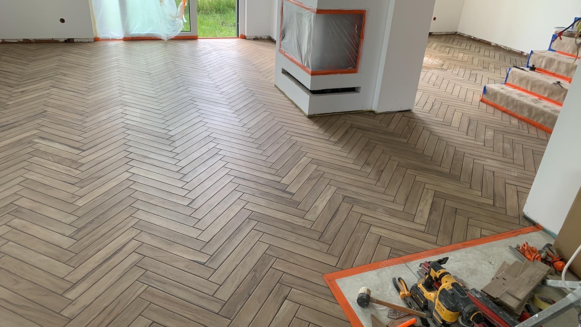 Ułożona podłoga z drewnopodobnych płytek w jodełkę; widoczne narzędzia glazurnicze na podłodze w rogu pomieszczenia, zabezpieczony kominek i schody.