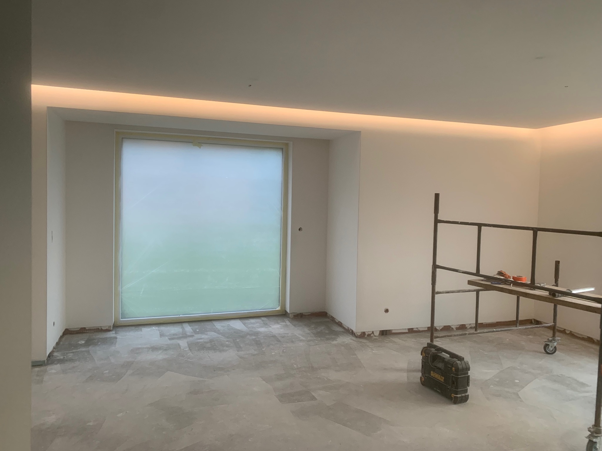 Pokój w trakcie remontu: okno zabezpieczone folią, ściany przygotowane do malowania z ukrytym oświetleniem LED, rozstawione rusztowanie i skrzynka narzędziowa firmy DeWalt.