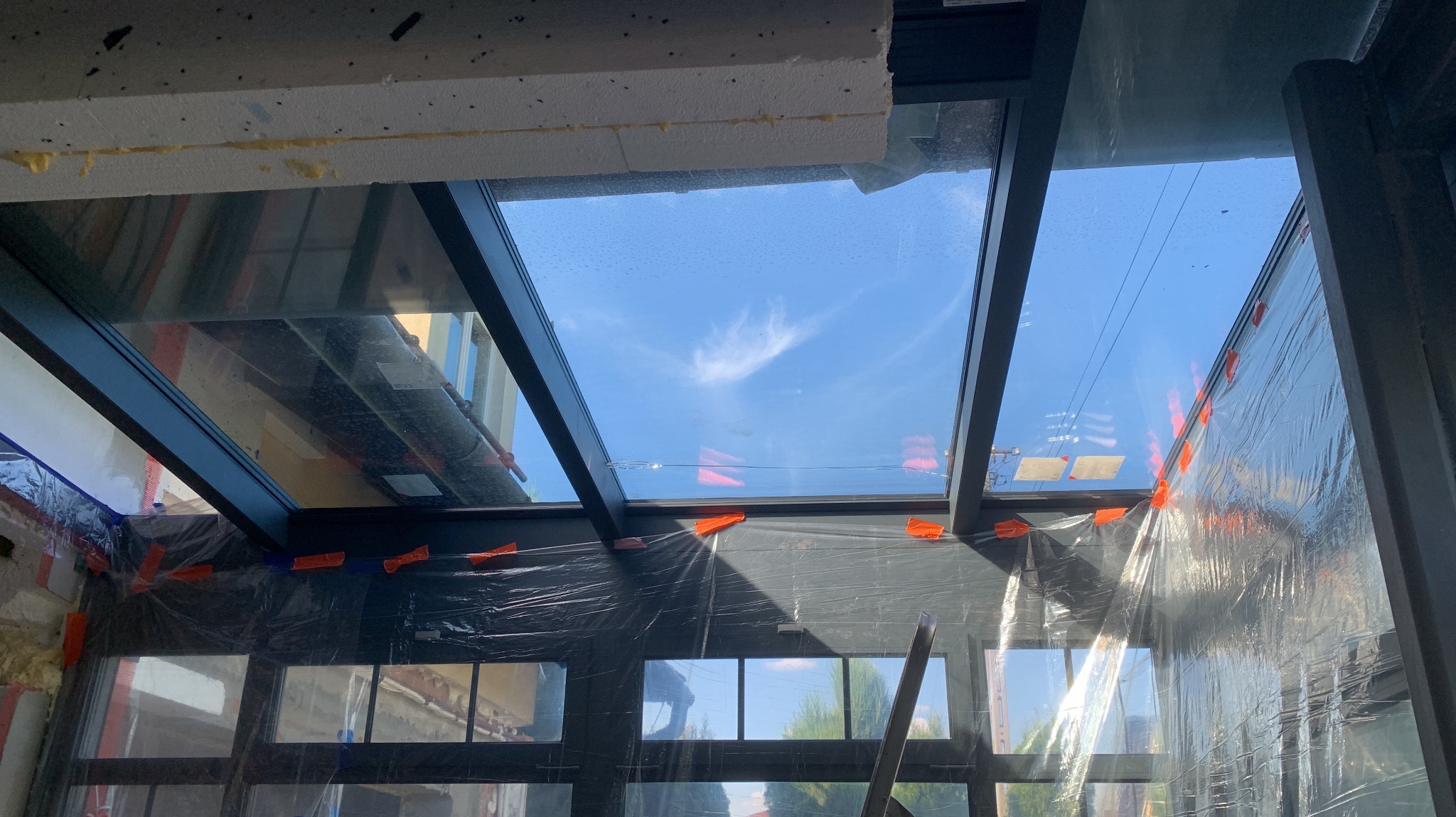 Szklany dach z czarną konstrukcją w trakcie montażu, zabezpieczony folią ochronną, widok na błękitne niebo z chmurami.