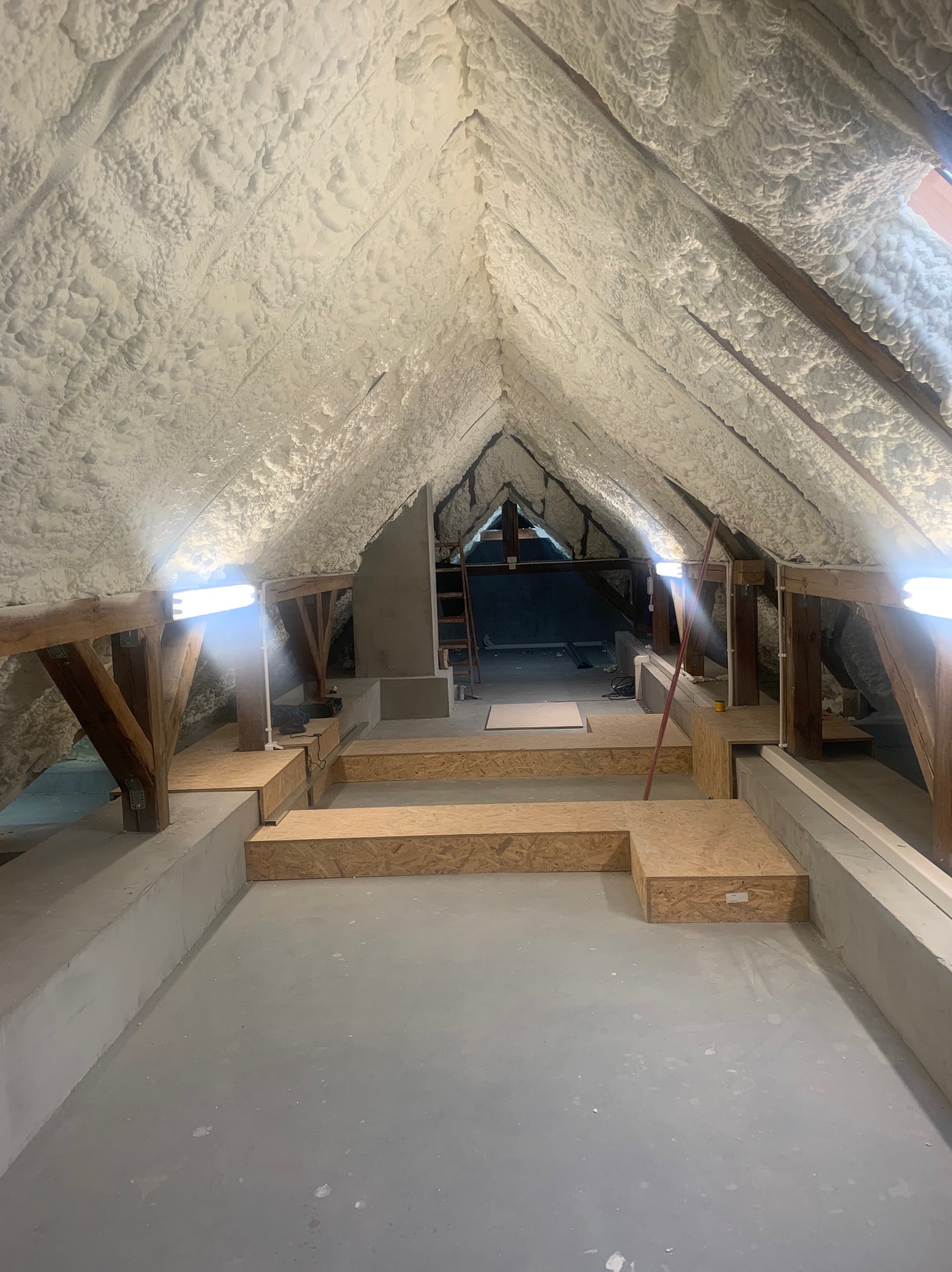 Poddasze ocieplone pianą PUR, widoczne drewniane belki konstrukcyjne, podłoga w trakcie przygotowania z płyt OSB, surowy beton na ścianach, oświetlenie halogenowe.