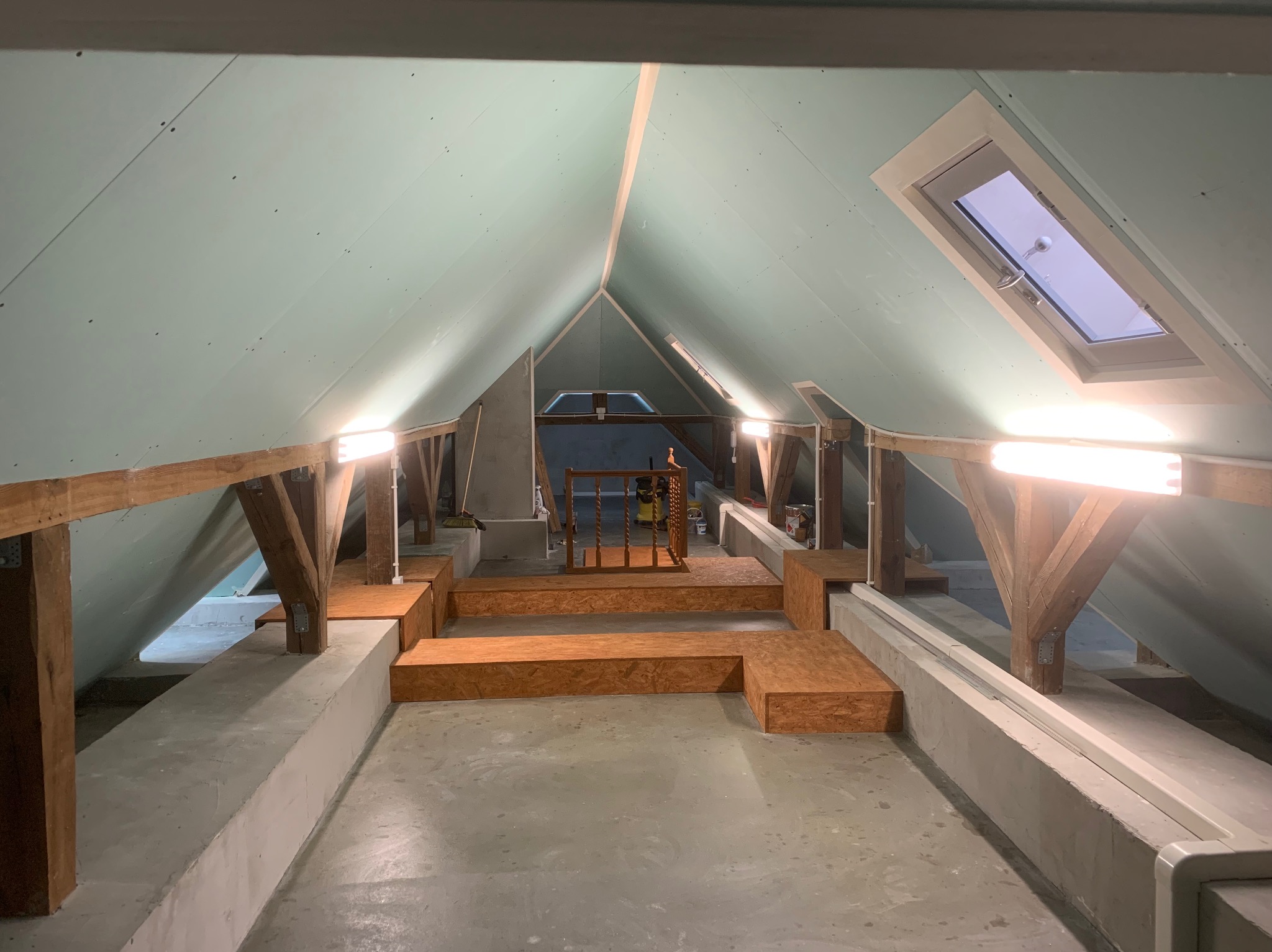 Poddasze w trakcie adaptacji: surowe ściany z płyt kartonowo-gipsowych, drewniane belki konstrukcyjne, okno dachowe i podest z płyty OSB, wstępny etap wykończenia.