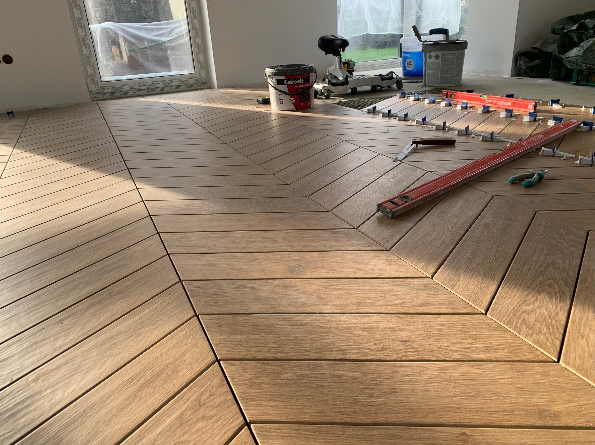 Ukończona podłoga z paneli ułożonych w jodełkę, z narzędziami glazurniczymi i poziomnicami. Widoczne klipsy do poziomowania. Jasne, naturalne światło.