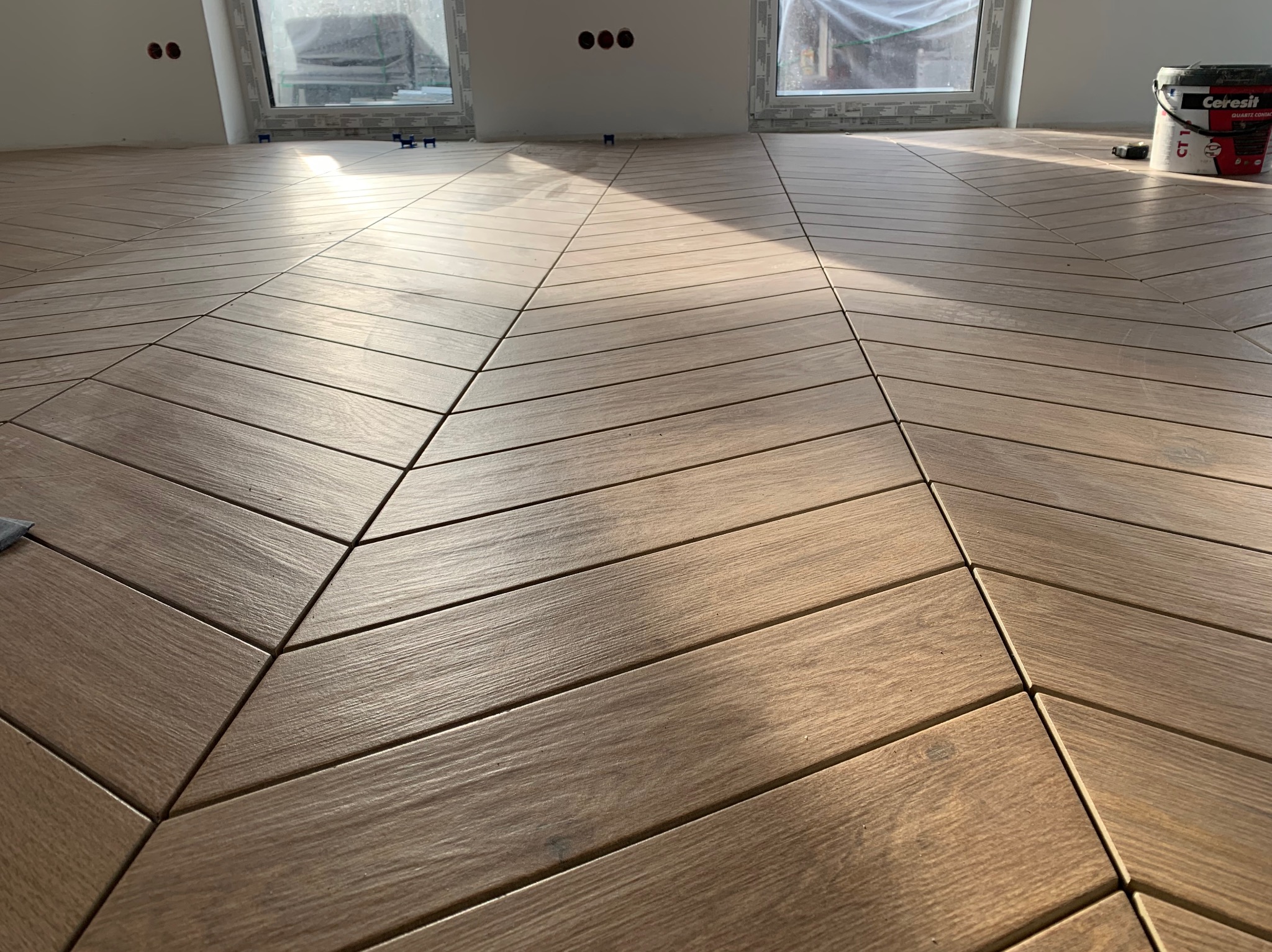 Ułożona podłoga z drewnopodobnych płytek w jodełkę, z widocznymi fugami i oknami w tle. Wiadro z klejem do płytek stoi w rogu pomieszczenia.