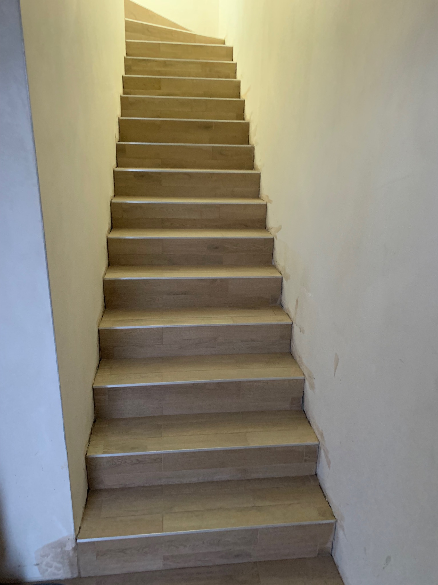 Wykończone schody wewnętrzne z drewnopodobnych płytek, prowadzące w górę. Widoczne niedoskonałości wykończenia ścian przy schodach. Ujęcie od dołu.