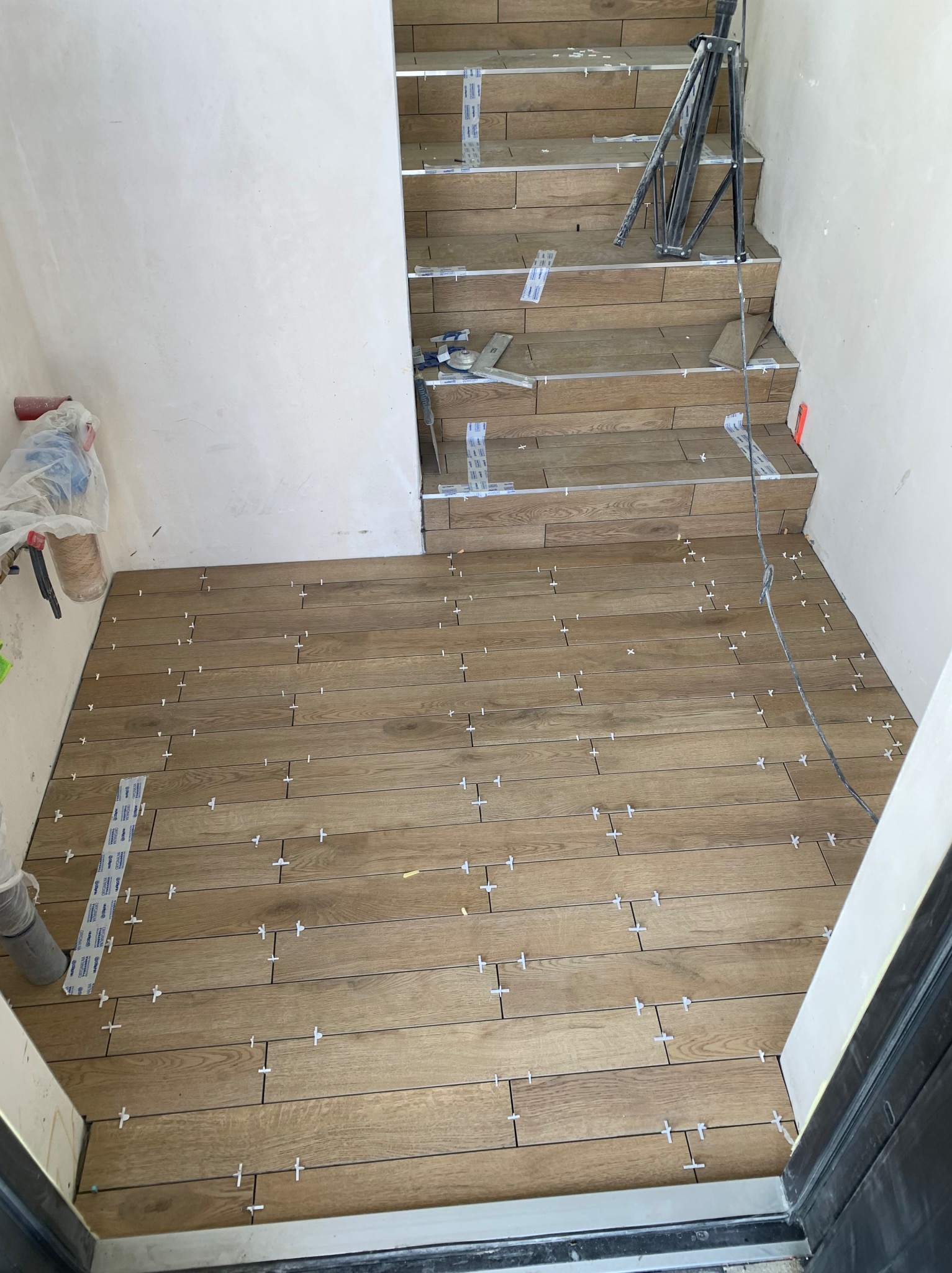 Układanie płytek imitujących drewno na podłodze i schodach. Widoczne krzyżyki dystansowe i narzędzia glazurnicze. Jasne ściany w tle.