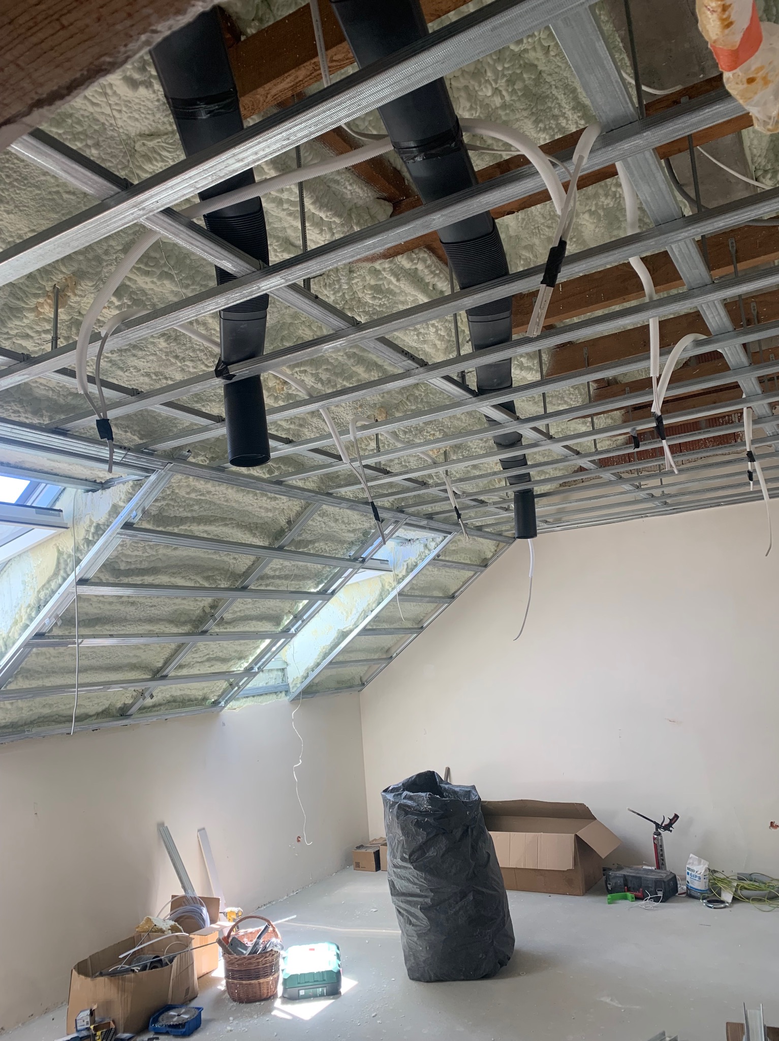 Poddasze w trakcie adaptacji: ocieplenie pianą, metalowy stelaż sufitu podwieszanego, instalacje. Widoczne narzędzia i materiały budowlane. Skos dachu z oknem.