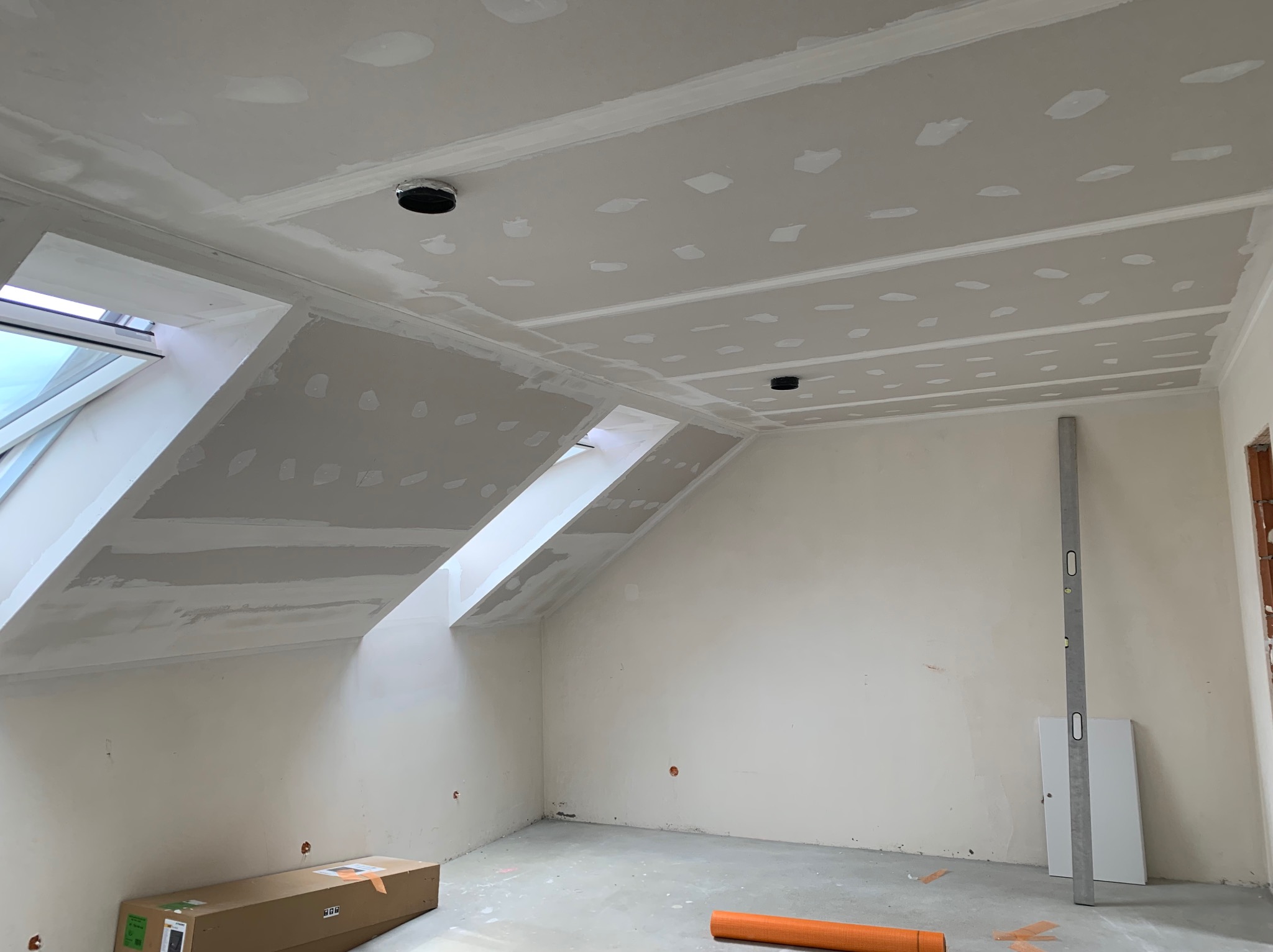 Poddasze z oknami dachowymi po montażu płyt gipsowo-kartonowych, widoczne szpachlowane łączenia, poziomica oparta o ścianę, przygotowane otwory na oświetlenie.