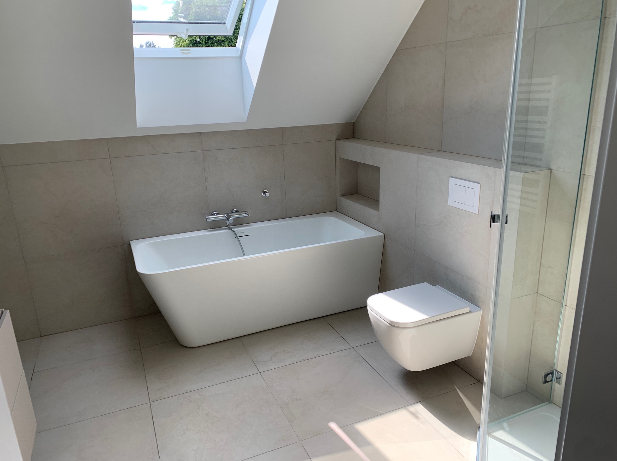 Jasna łazienka na poddaszu z wanną, toaletą podwieszaną i kabiną prysznicową. Beżowe płytki, okno dachowe. Minimalistyczny design.