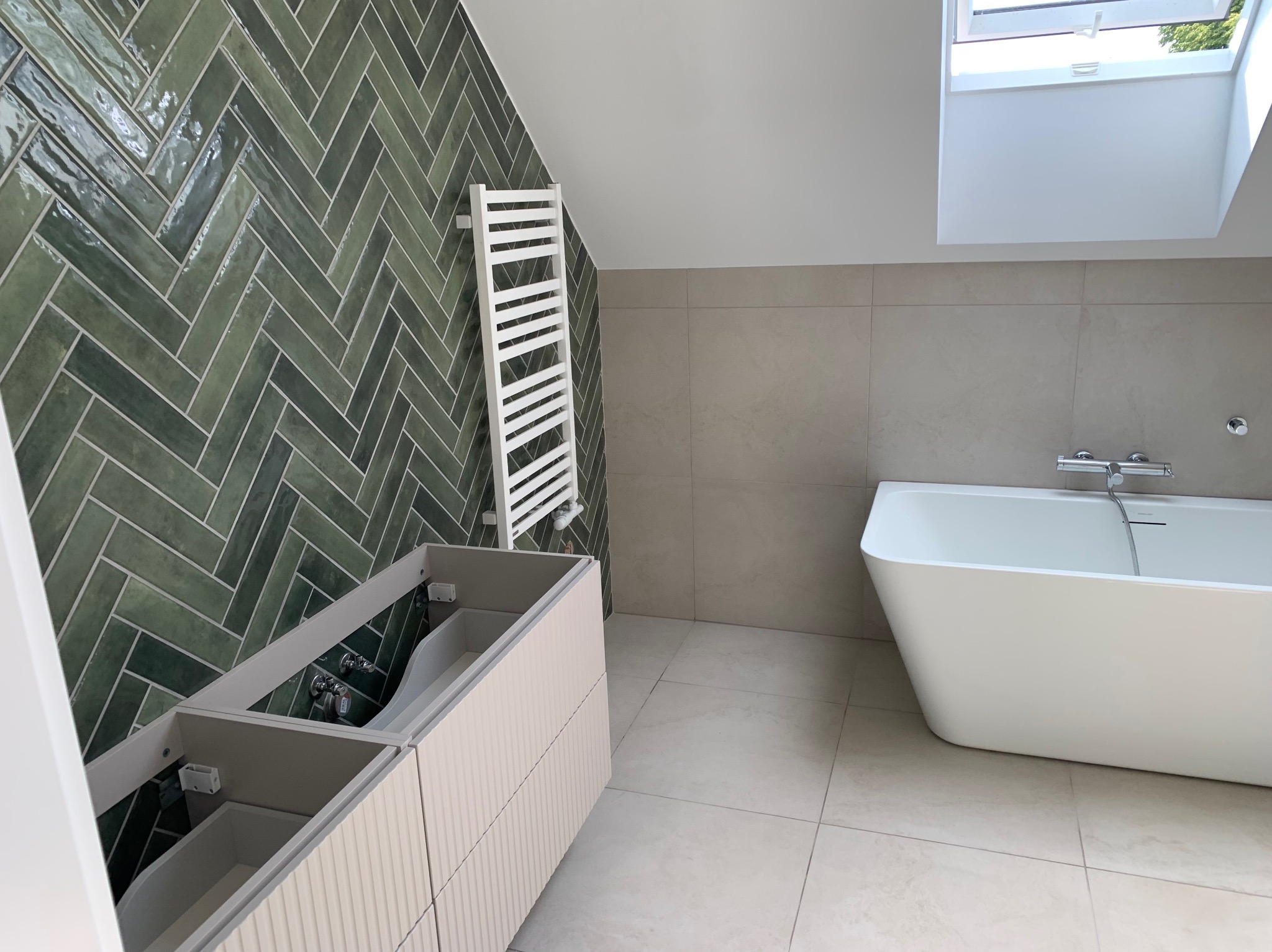 Nowoczesna łazienka w skosach: zielona ściana z płytek ułożonych w jodełkę, biała wanna wolnostojąca, grzejnik drabinkowy i szafka pod umywalkę w trakcie montażu.