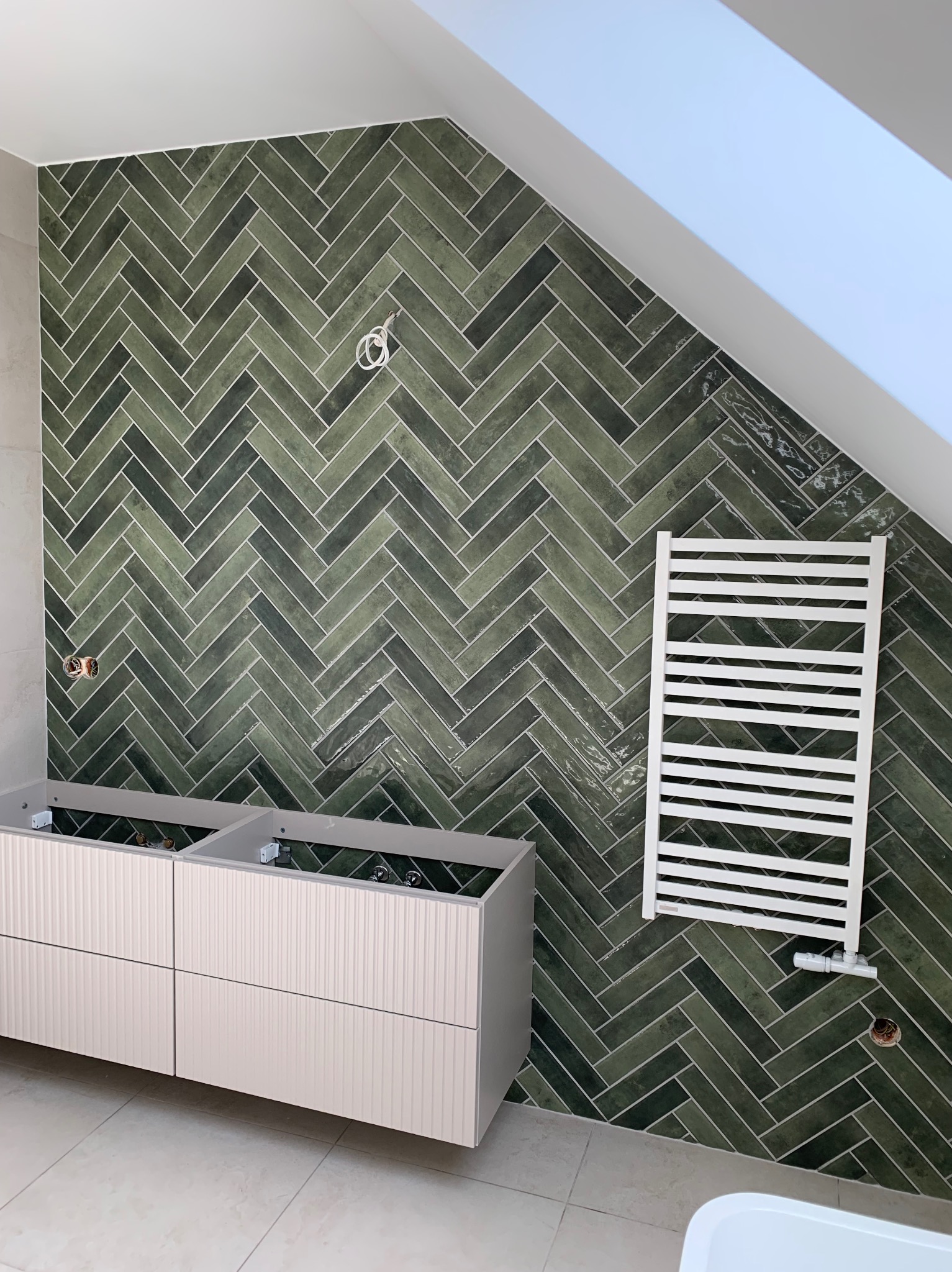 Ukończona ściana łazienki z zielonej, błyszczącej glazury ułożonej w jodełkę. Widoczny grzejnik drabinkowy, szafka pod umywalkę i fragment okna dachowego.