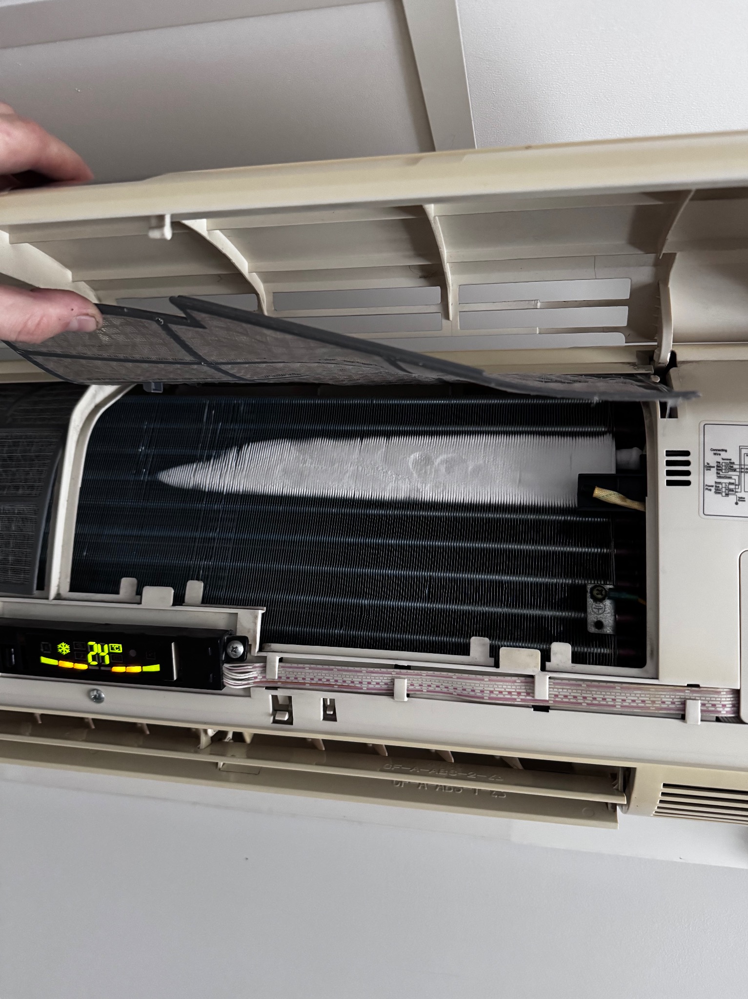 Otwarty panel klimatyzatora z widocznym filtrem i żebrami chłodzącymi, wyświetlacz pokazuje 24 stopnie Celsjusza, widoczna dłoń przytrzymująca panel.