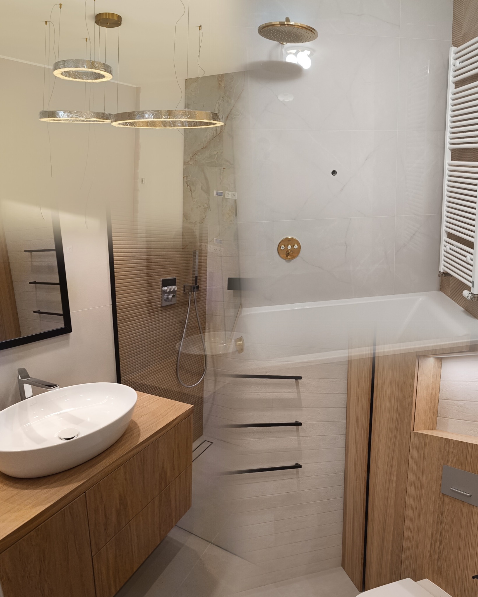 Nowoczesna łazienka z drewnianą szafką pod umywalką, białymi płytkami imitującymi marmur w strefie prysznicowej z deszczownicą i grzejnikiem drabinkowym, oraz okrągłymi lampami wiszącymi.