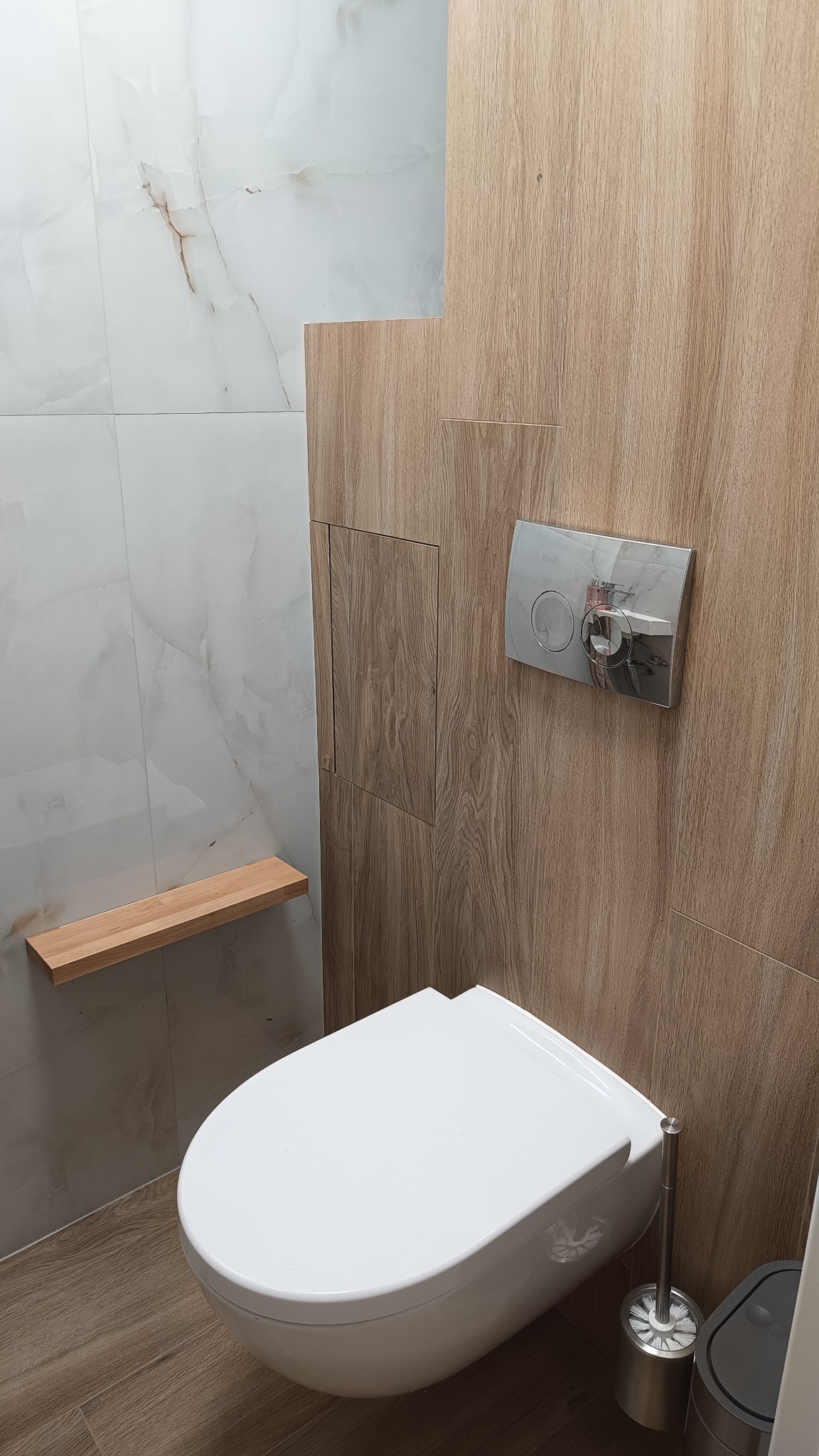 Nowoczesna toaleta podwieszana z ukrytą spłuczką, drewnopodobne panele ścienne i marmurowe płytki, drewniana półka nad toaletą.