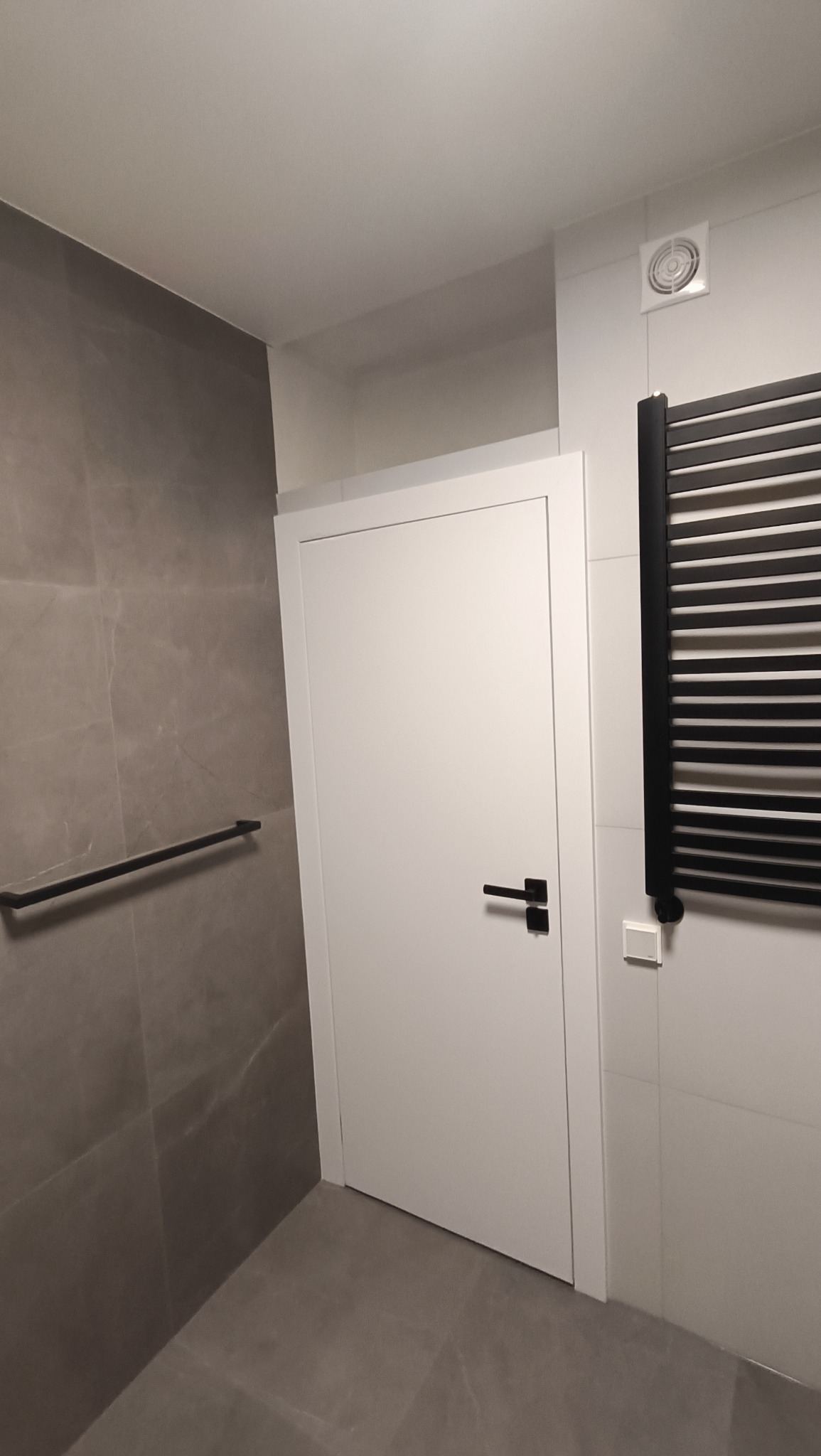 Nowoczesna łazienka z szarymi płytkami imitującymi marmur, białe drzwi z czarną klamką, czarny grzejnik drabinkowy i czarny reling na ręczniki.