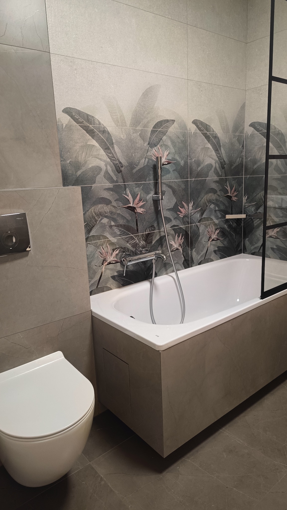 Nowoczesna łazienka z wanną obudowaną płytkami imitującymi beton, toaletą wiszącą i płytkami ściennymi z motywem tropikalnych liści i kwiatów.
