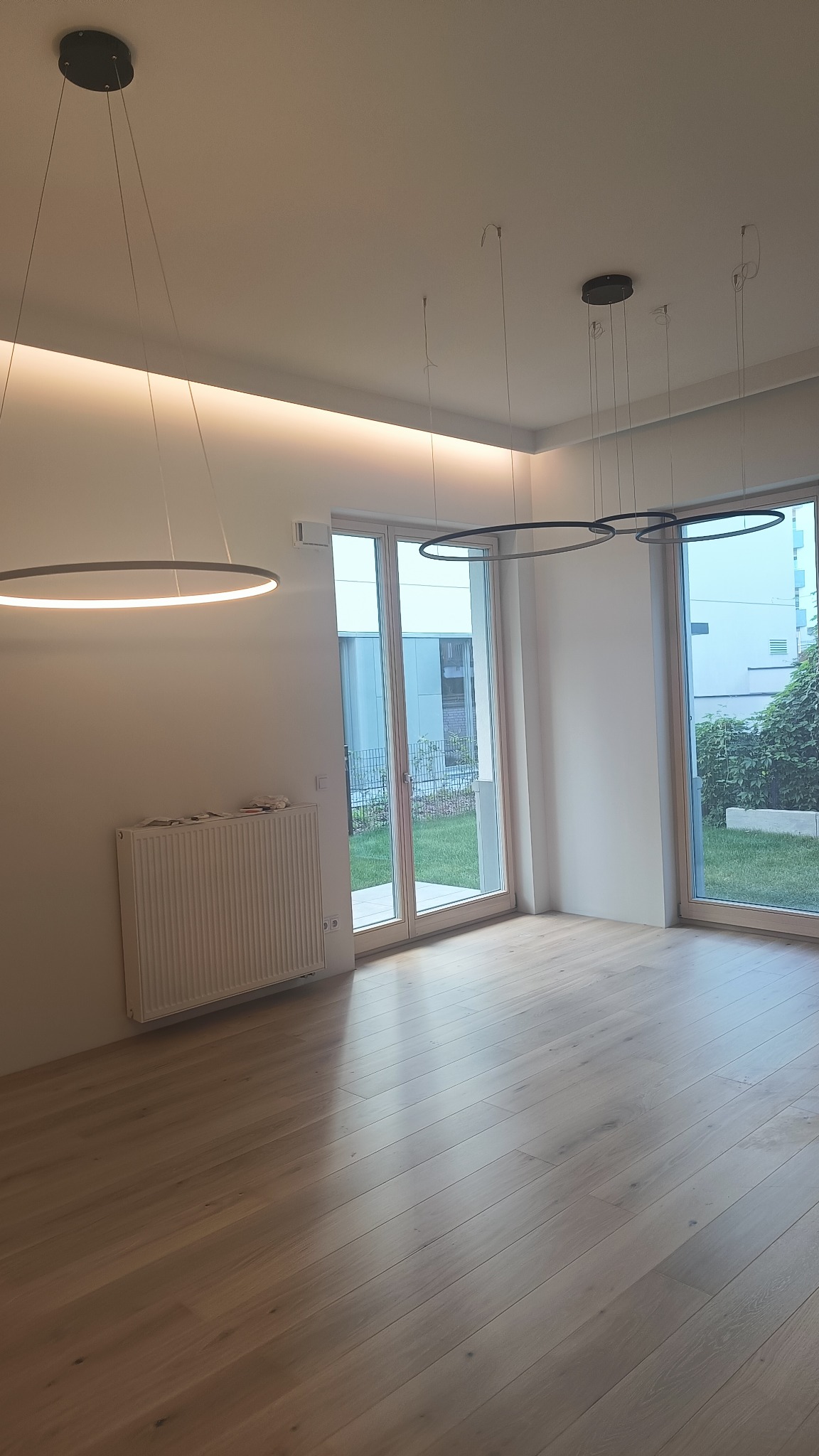 Puste, jasne wnętrze z drewnianą podłogą, białymi ścianami, grzejnikiem i trzema nowoczesnymi, okrągłymi lampami wiszącymi. Widoczne duże okna balkonowe z widokiem na ogród.