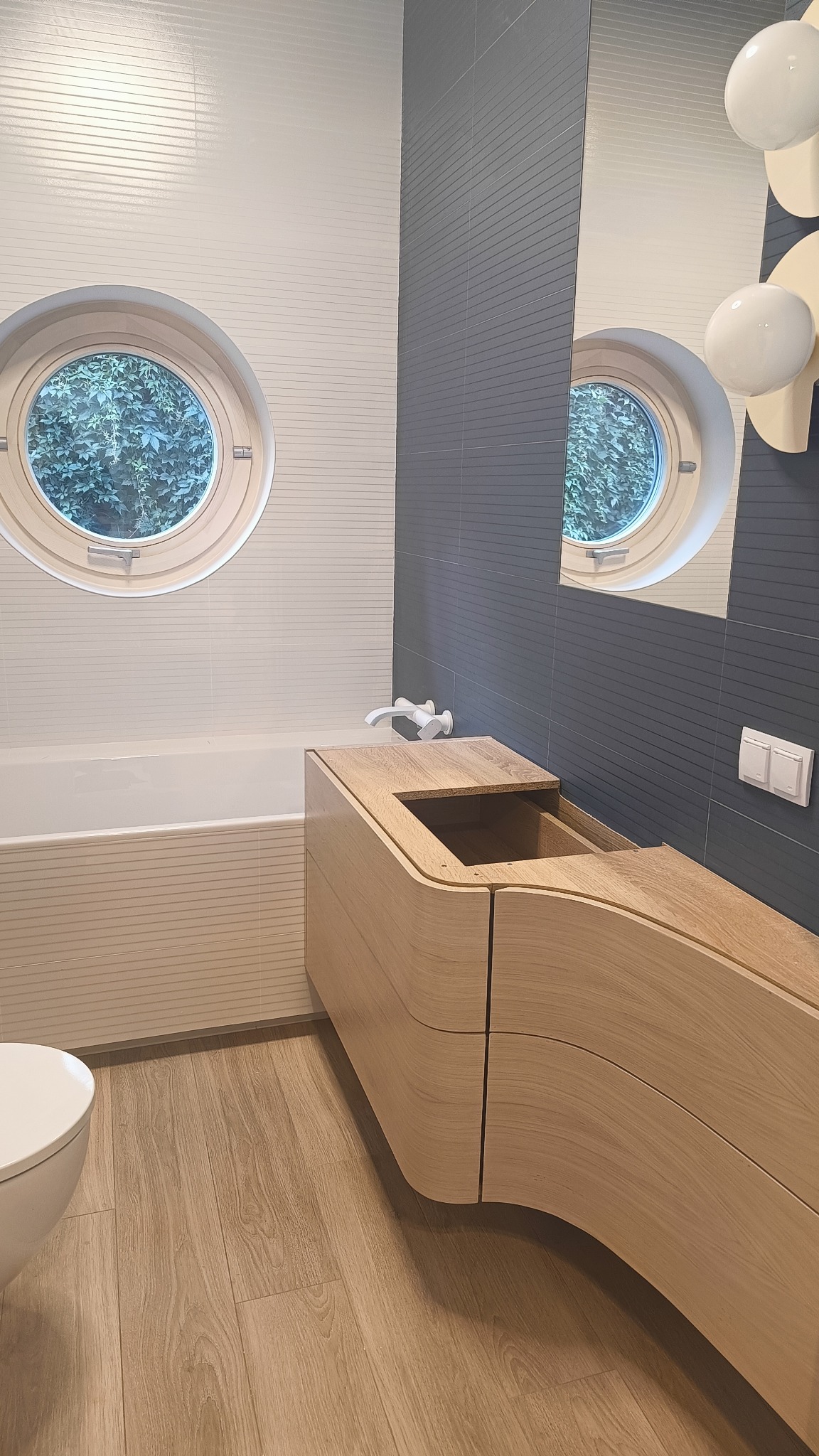 Nowoczesna łazienka z okrągłym oknem, drewnianą szafką pod umywalką o zaokrąglonych kształtach, szarą ścianą z poziomymi płytkami oraz podłogą imitującą drewno.