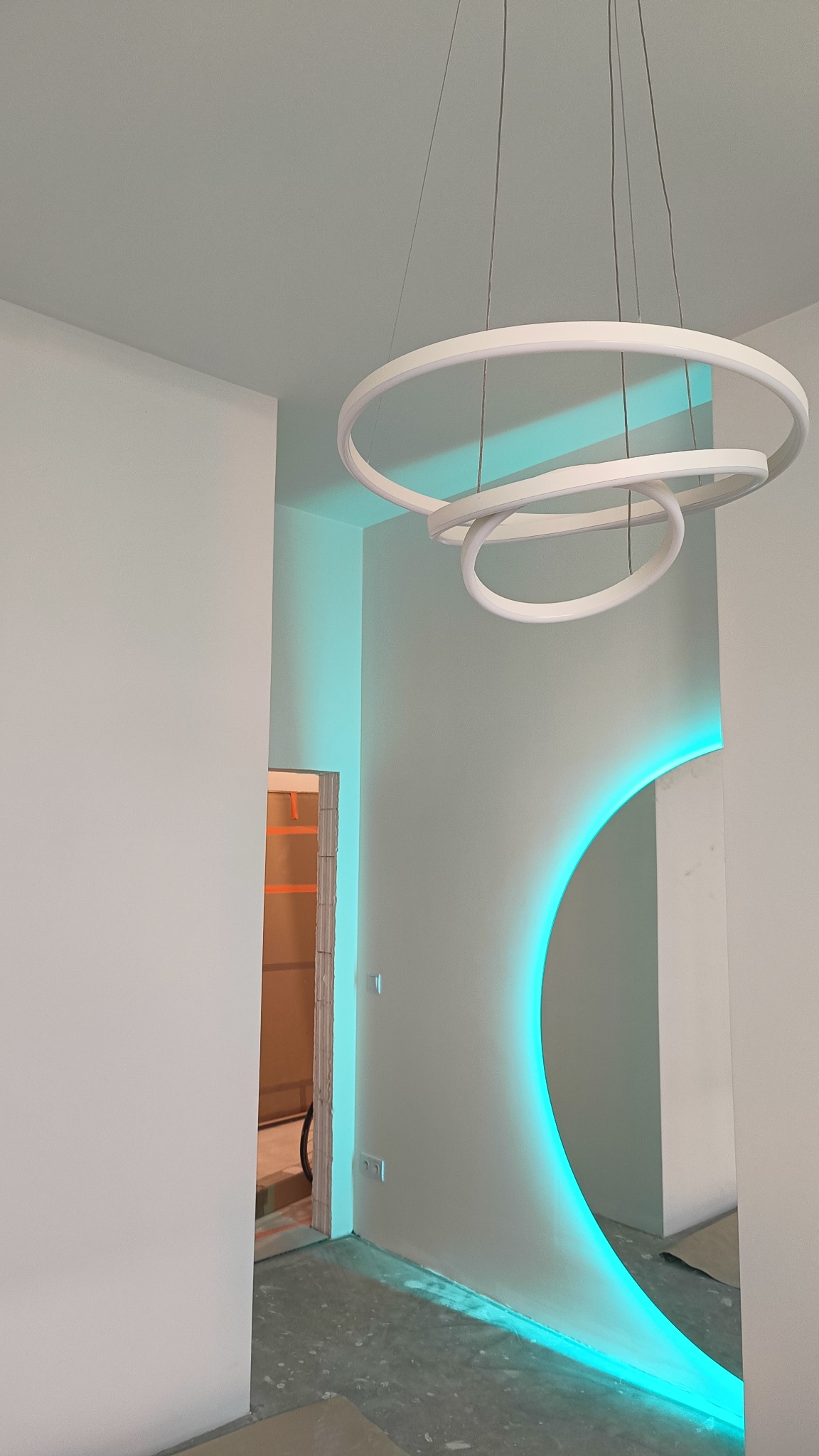 Nowoczesne wnętrze z podświetlonym na niebiesko łukiem w ścianie, wiszącą lampą o kształcie trzech połączonych okręgów i widocznym fragmentem drzwi z zabezpieczeniami.