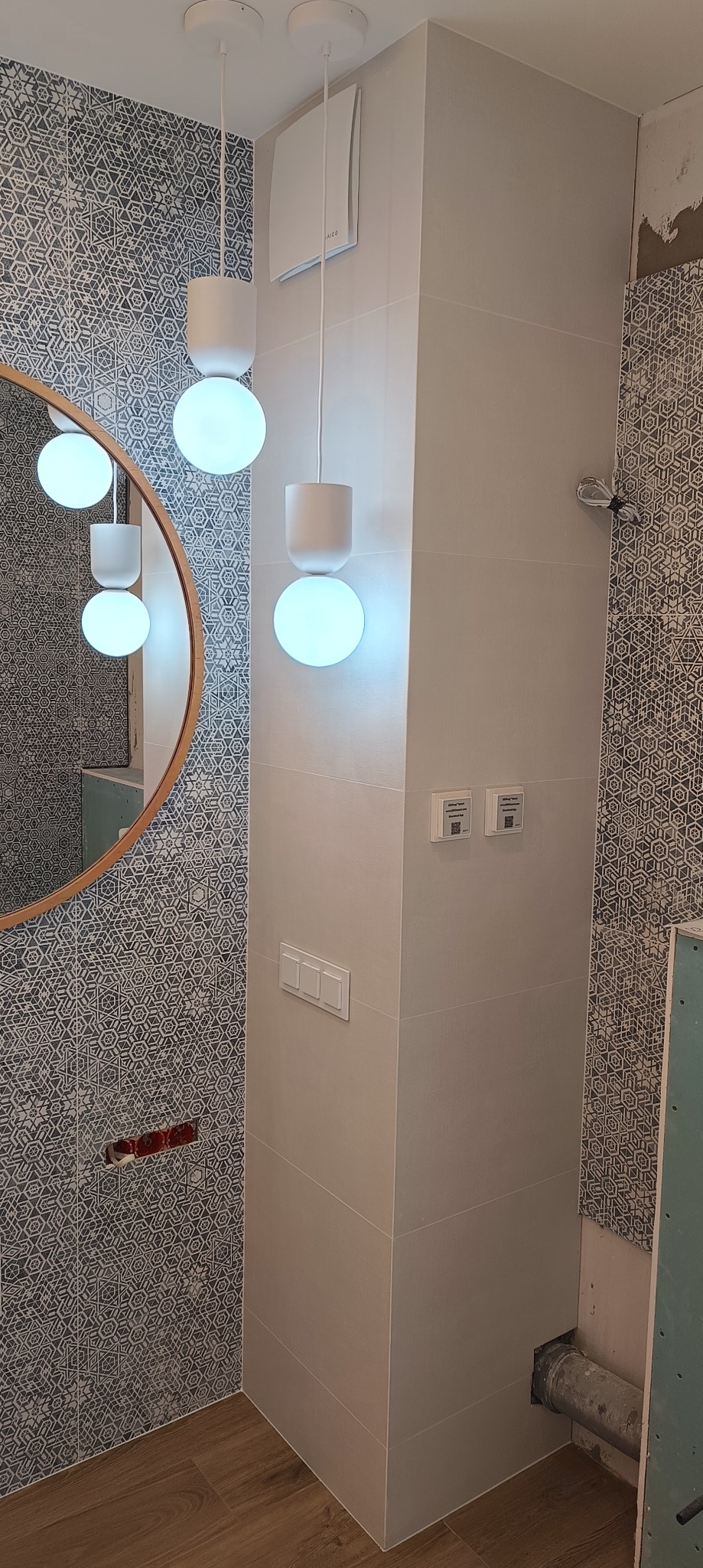 Fragment łazienki w trakcie wykańczania: dekoracyjne płytki ścienne w geometryczny wzór, drewnopodobna podłoga, kolumna z jasnych płytek z włącznikami światła i trzema okrągłymi lampami wiszącymi...