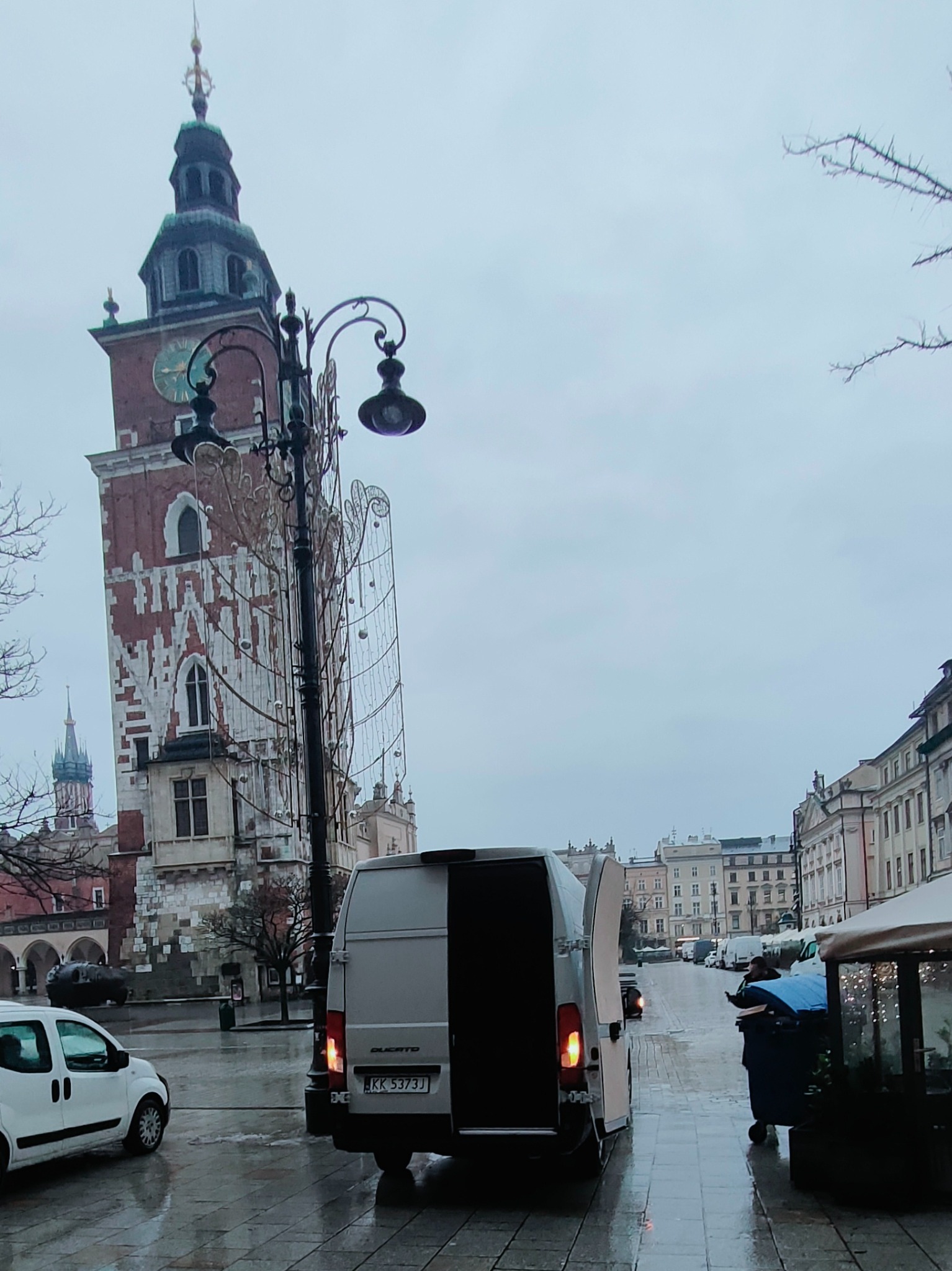 Szary bus z otwartymi drzwiami z tyłu, zaparkowany na mokrym Rynku Głównym w Krakowie, na tle Sukiennic i wieży Ratuszowej w pochmurny dzień.