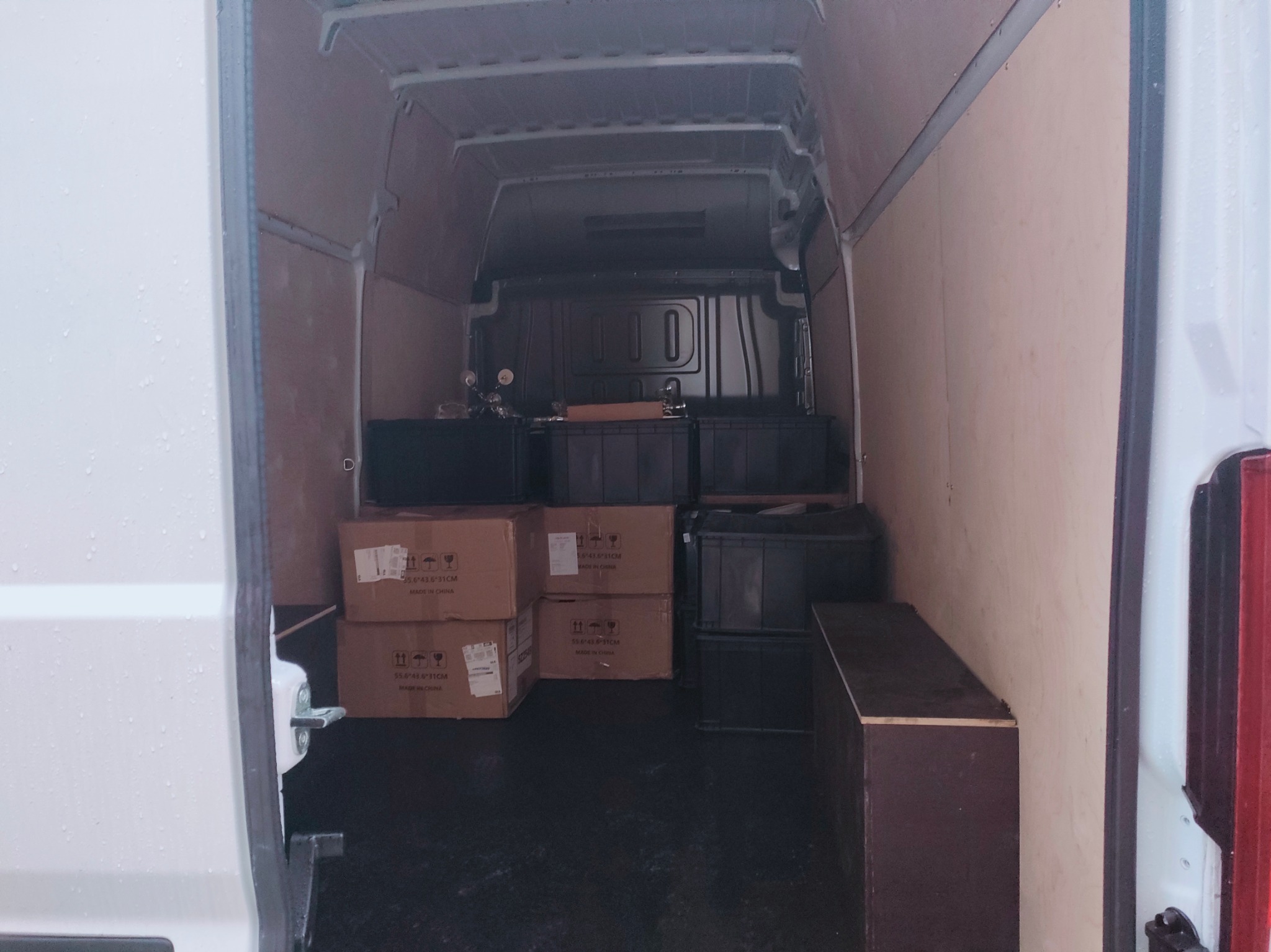 Wnętrze furgonetki z częściowo załadowanymi kartonami i czarnymi skrzynkami, widok z tyłu pojazdu.