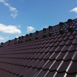 USŁUGI DEKARSKIE IRENEUSZ SUWAŁA - Brązowy dach z blachy trapezowej z rzędem czarnych kominków wentylacyjnych na tle błękitnego nieba z chmurami, widoczne elementy konstrukcji drewnianej.