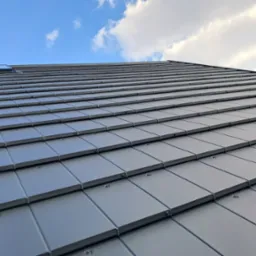 Szary dach z dachówki ceramicznej widziany pod kątem, z oknem dachowym i fragmentem nieba z chmurami w tle. Widoczne mocowania dachówek.