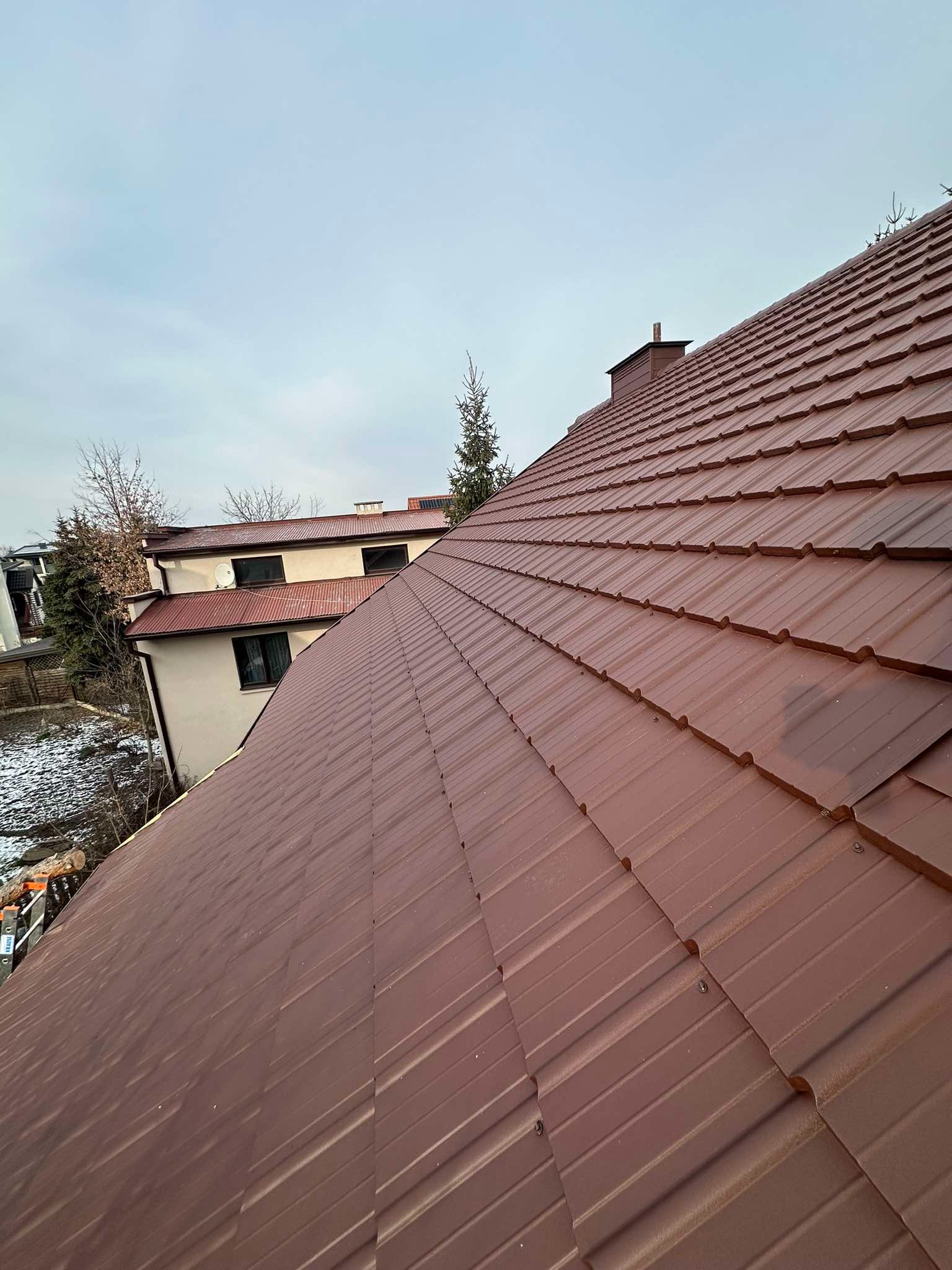 Widok z góry na nowo położony, brązowy dach z blachy trapezowej na tle zabudowań i pochmurnego nieba. Widoczne kominy i fragment drabiny na skraju dachu.