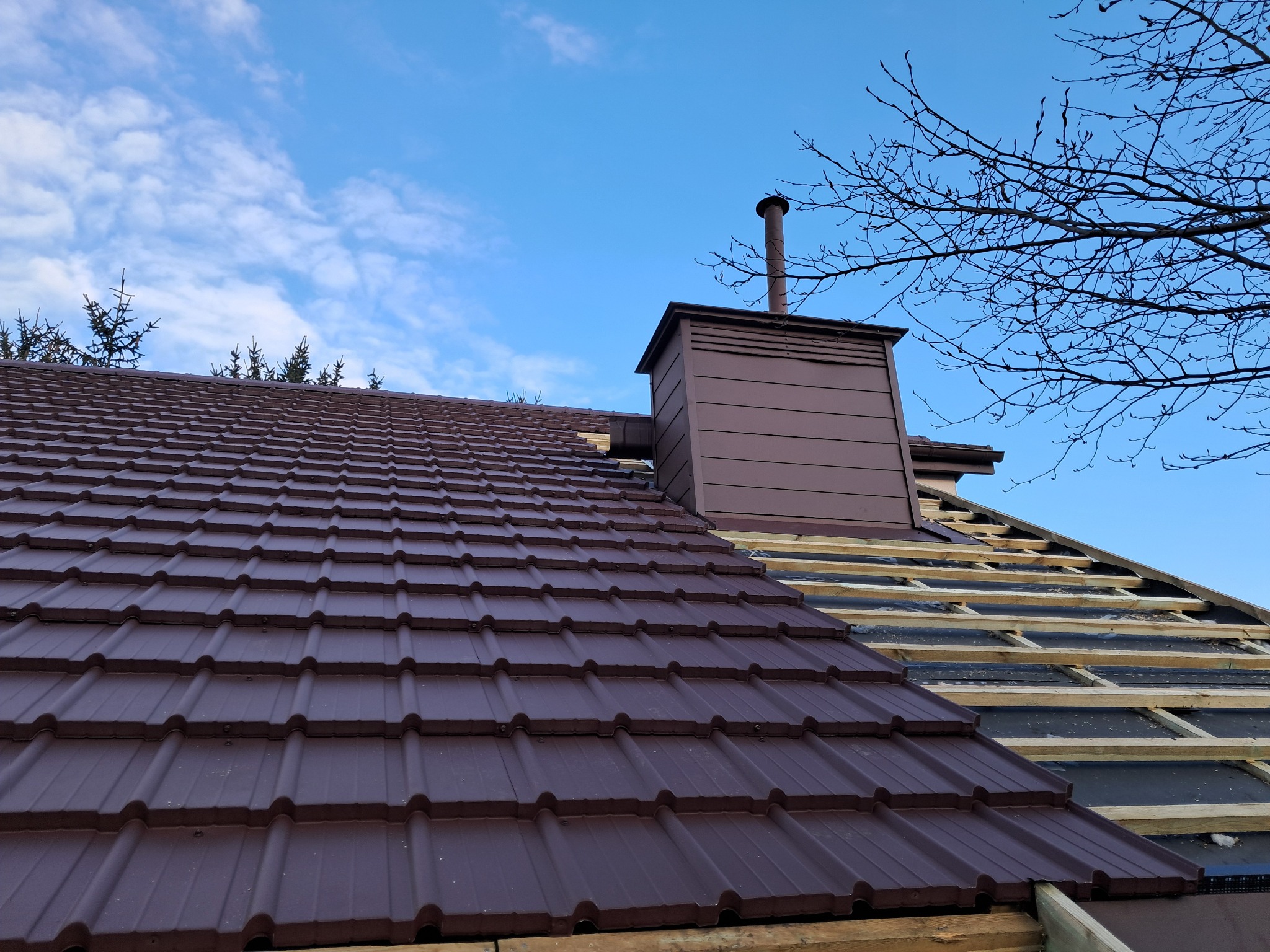 Częściowo pokryty dach z brązową dachówką, widoczny komin z metalowym wykończeniem oraz fragment drewnianej konstrukcji dachu w trakcie prac, na tle błękitnego nieba i gałęzi drzewa.