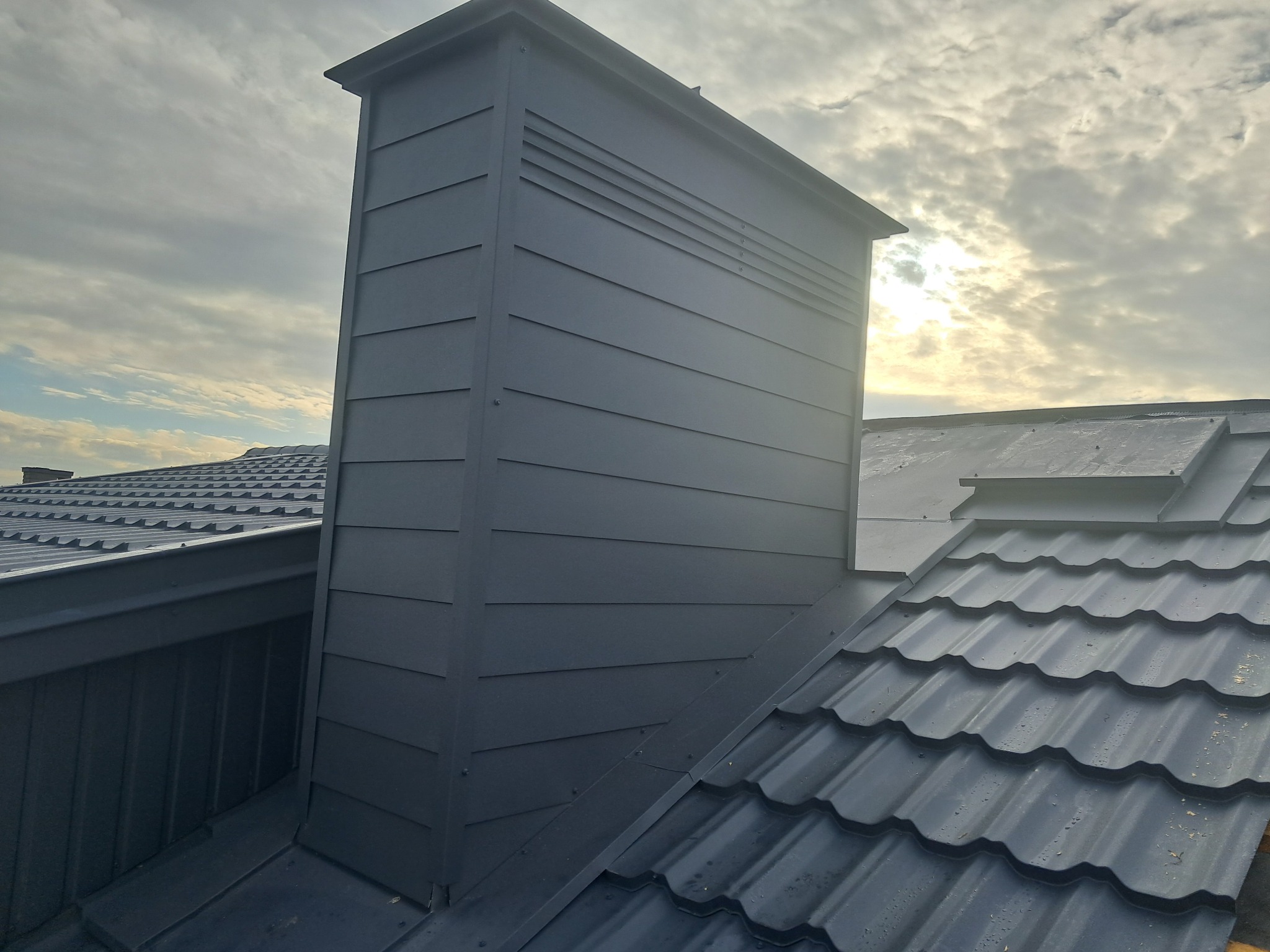 Szary komin wentylacyjny z poziomymi żaluzjami na dachu pokrytym blachodachówką modułową w odcieniach szarości, na tle zachmurzonego nieba z przebijającym słońcem.
