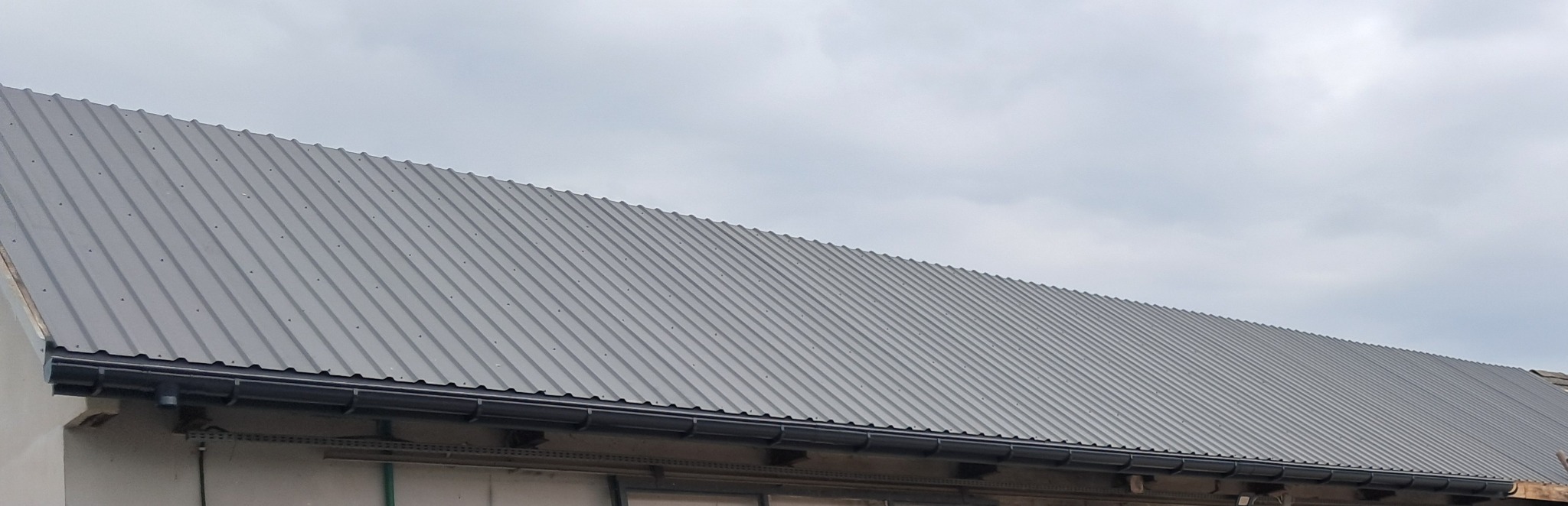 Szary dach z blachy trapezowej z zamontowanymi czarnymi rynnami, widok z dołu na tle pochmurnego nieba.