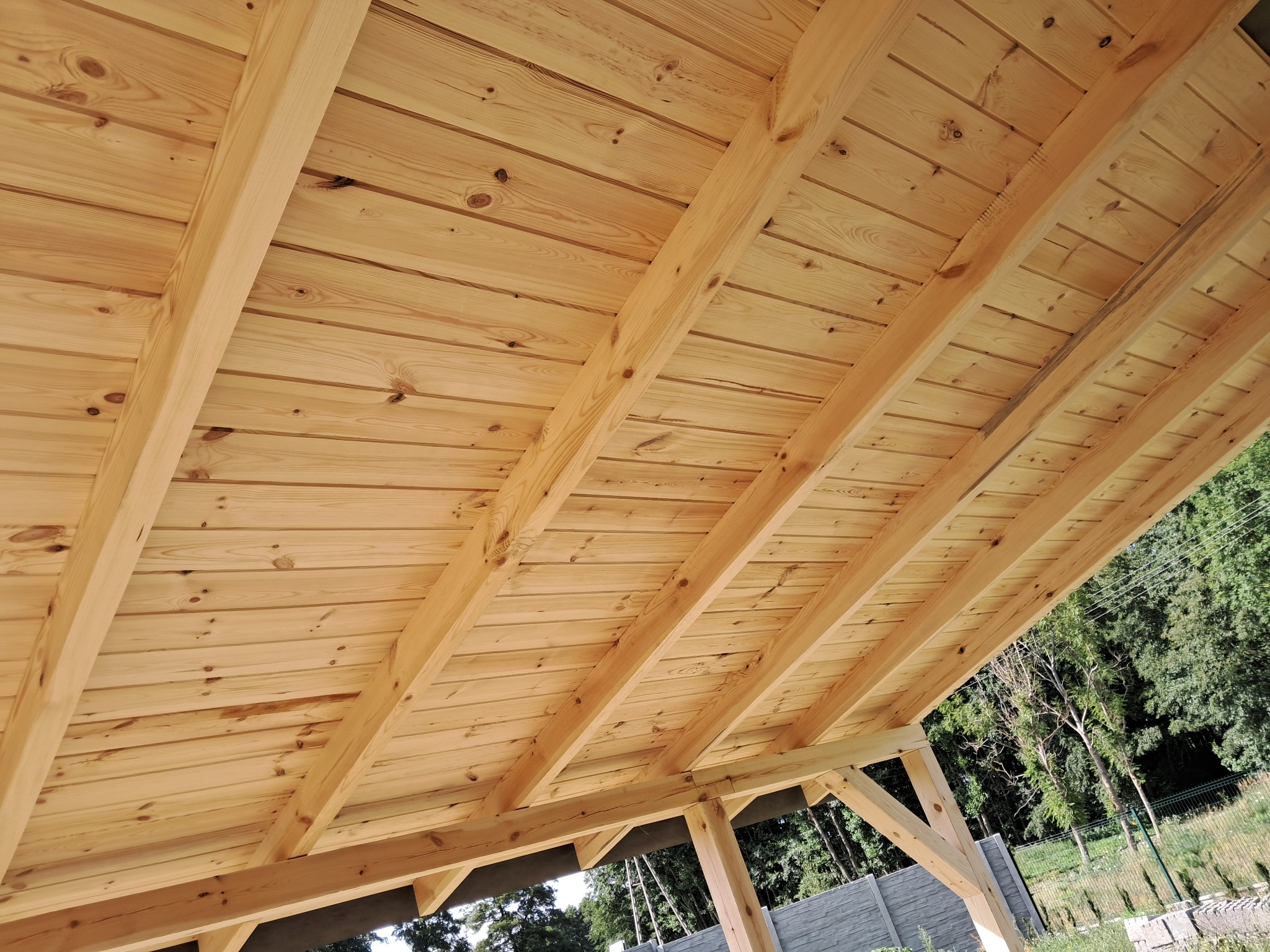 Drewniany strop z widocznymi belkami nośnymi, wykonany z jasnego drewna, z widocznymi słojami i sękami, na tle zieleni drzew i betonowego ogrodzenia.
