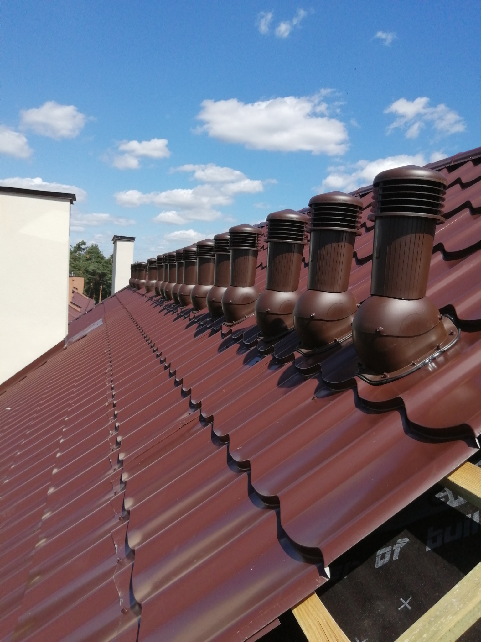 Nowy dach z brązowej dachówki z zamontowanymi kominkami wentylacyjnymi w rzędzie, widok z dołu na błękitne niebo z chmurami.