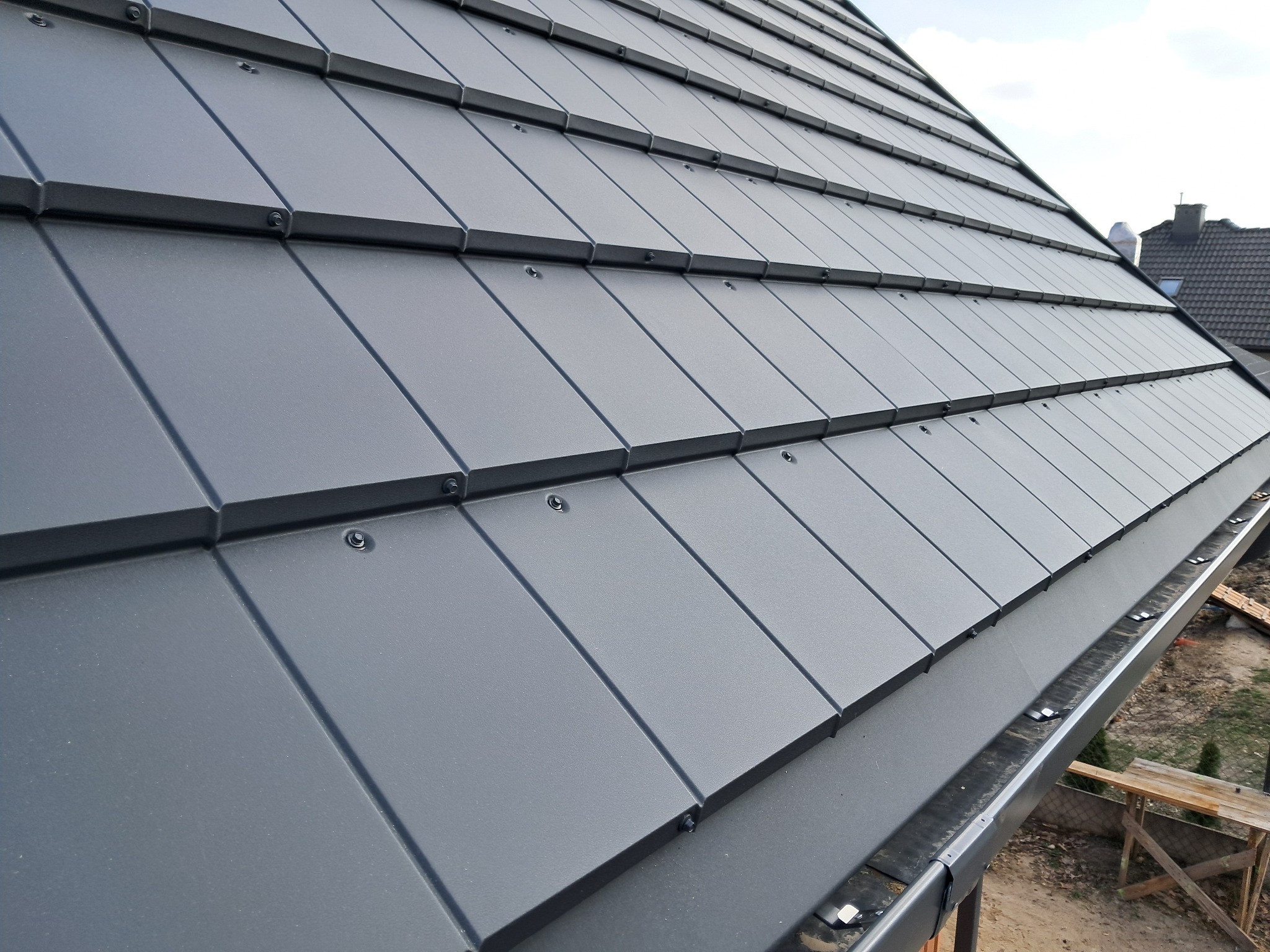 Szary dach z blachy trapezowej, widok z dołu, z zamontowanymi rynnami. Widoczne elementy mocujące i faktura blachy.