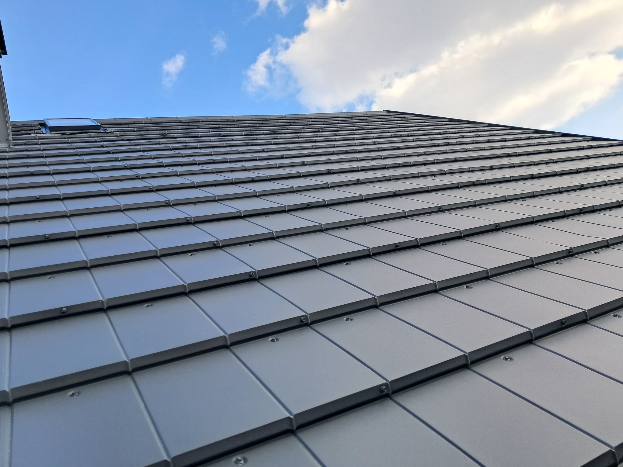 Szary dach z dachówki ceramicznej widziany pod kątem, z oknem dachowym i fragmentem nieba z chmurami w tle. Widoczne mocowania dachówek.