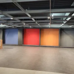 Wnętrze komercyjne z pomalowanymi na różne kolory ścianami (niebieski, czerwony, pomarańczowy). Widoczna szara podłoga i drewniane meble. Oświetlenie sufitowe.