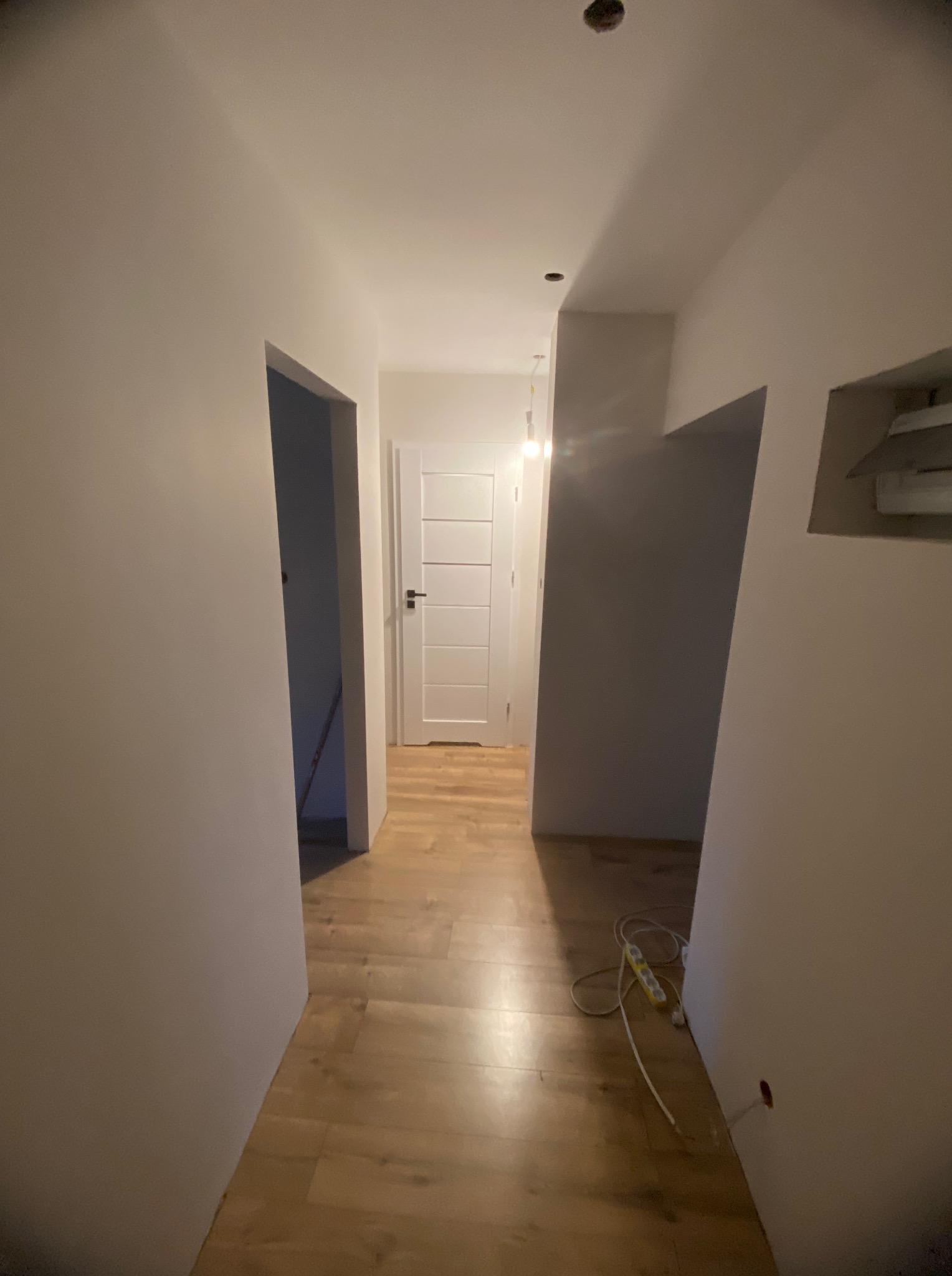 Korytarz po remoncie: gładkie, pomalowane ściany w odcieniach bieli, drewniana podłoga, białe drzwi. Widoczna listwa zasilająca przy ścianie.