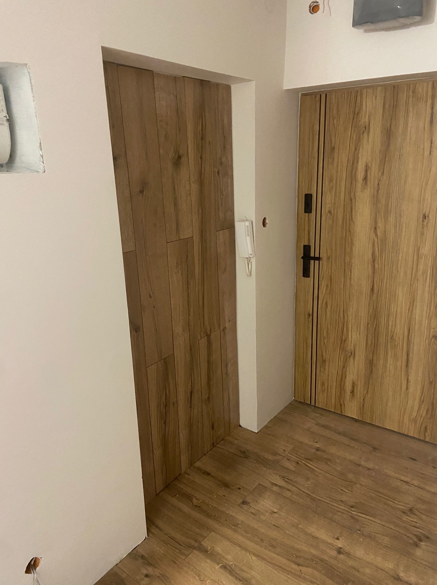 Wykończone wnętrze: drewnopodobne panele na podłodze i drzwiach, świeżo pomalowane ściany na biało. Widoczne detale wykończeniowe, takie jak listwy przypodłogowe i ościeżnice.