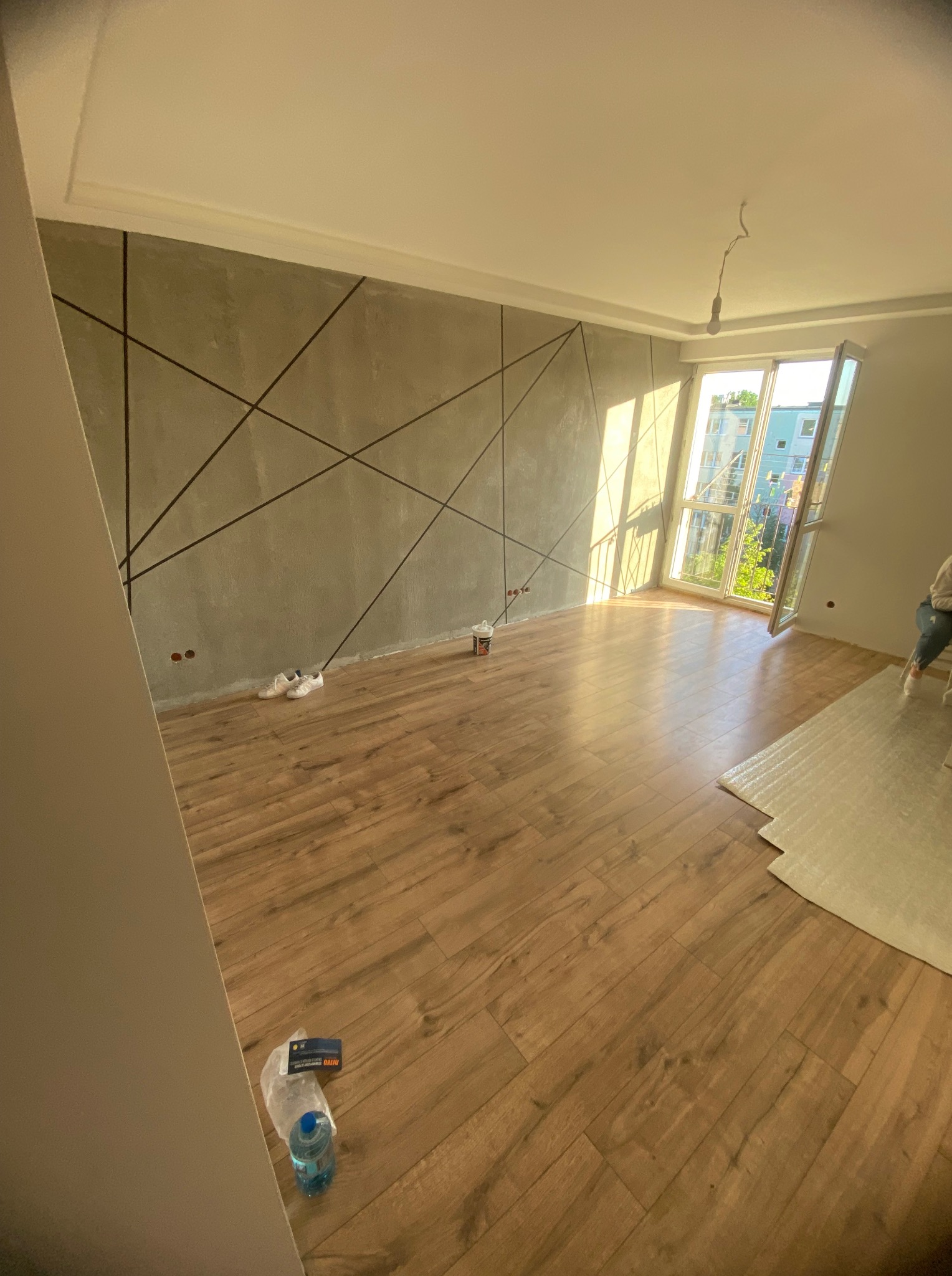 Jasne wnętrze po remoncie: drewnopodobne panele, szara ściana z geometrycznym wzorem, wpadające promienie słońca przez otwarte okno.