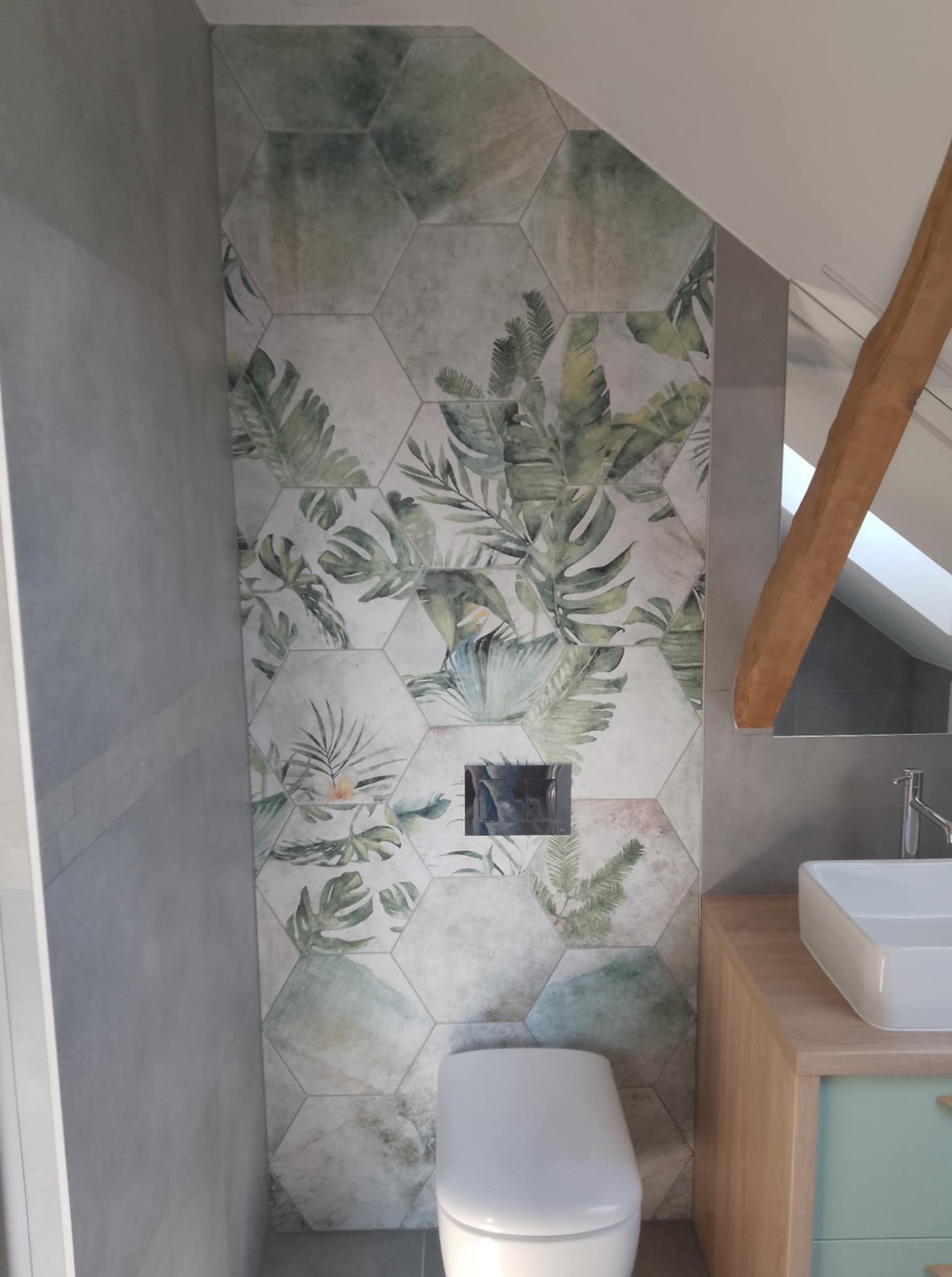 Wykończona łazienka na poddaszu z heksagonalnymi płytkami w tropikalne wzory na ścianie, umywalką nablatową i widoczną drewnianą belką stropową.