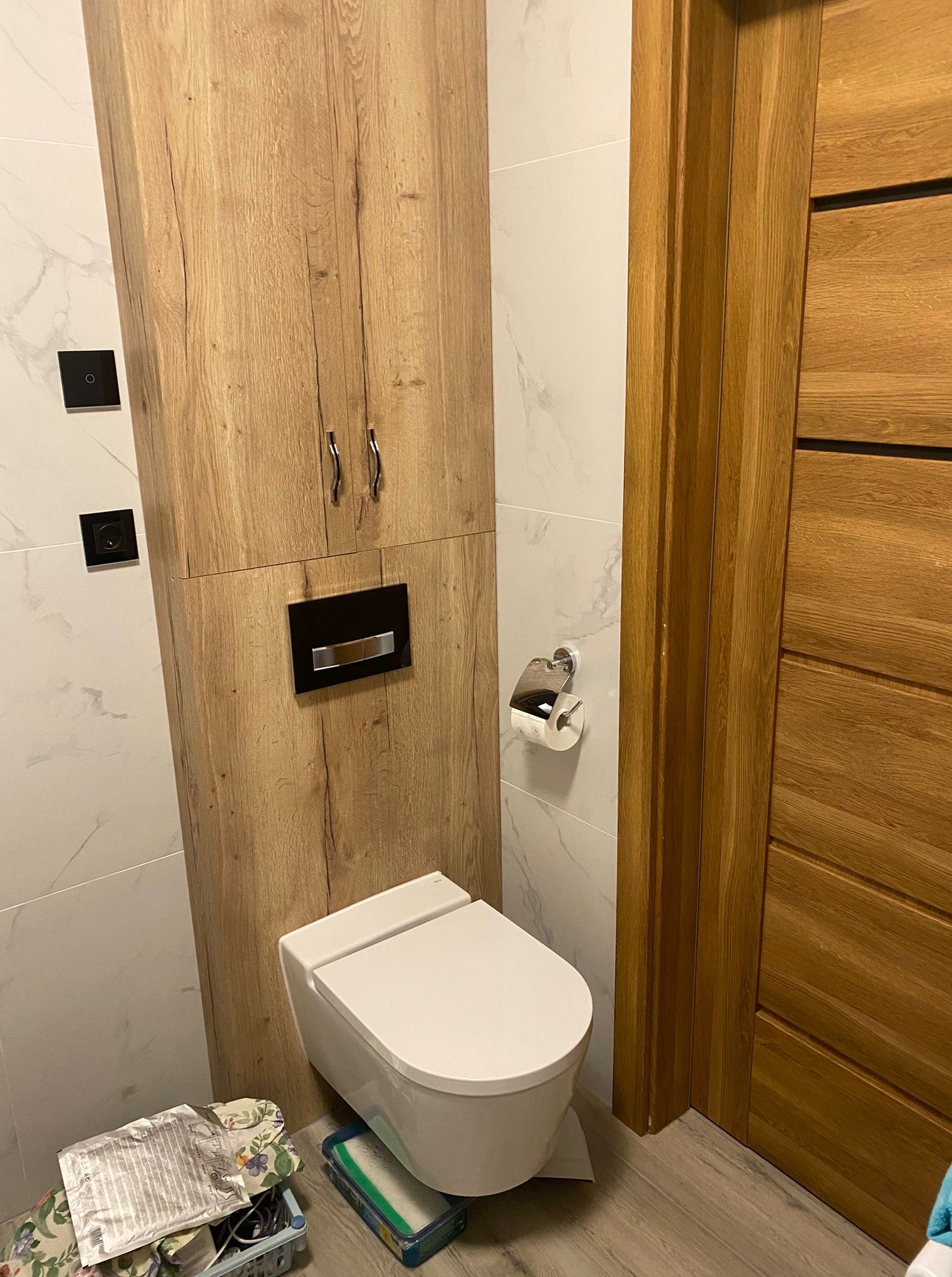 Nowoczesna toaleta z drewnianą szafką i drzwiami, marmurowe płytki, czarne włączniki, minimalistyczny styl. Widoczne akcesoria toaletowe i fragment podłogi.