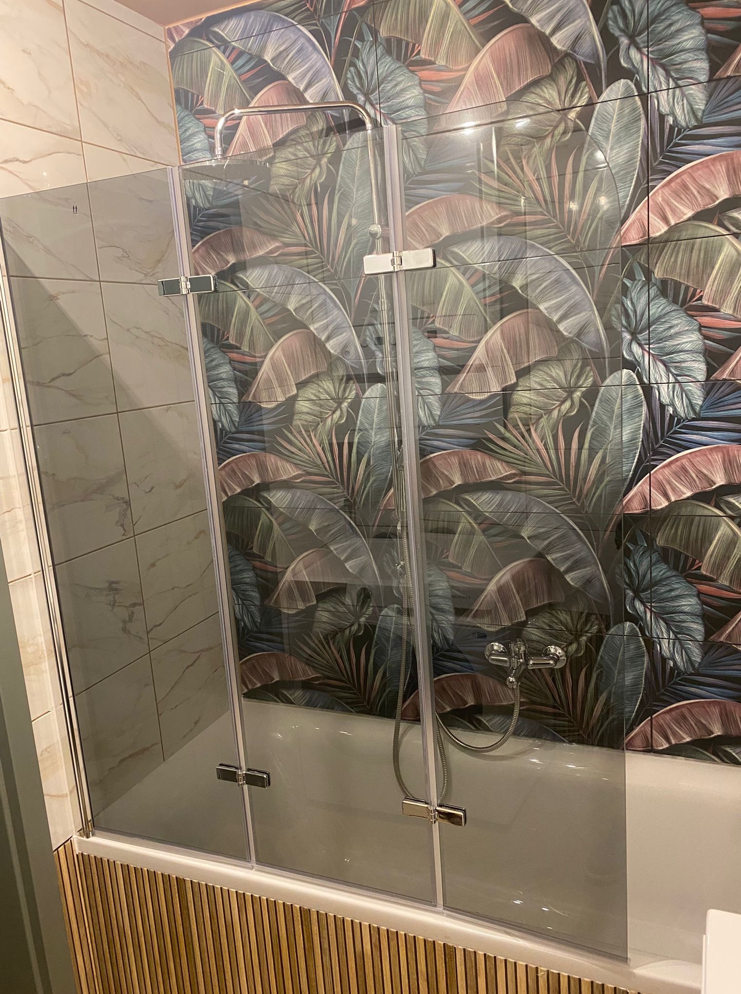 Wykończona łazienka z kabiną nawannową, płytkami imitującymi marmur i wzorem liści. Widoczna armatura i drewnopodobne panele na obudowie wanny.