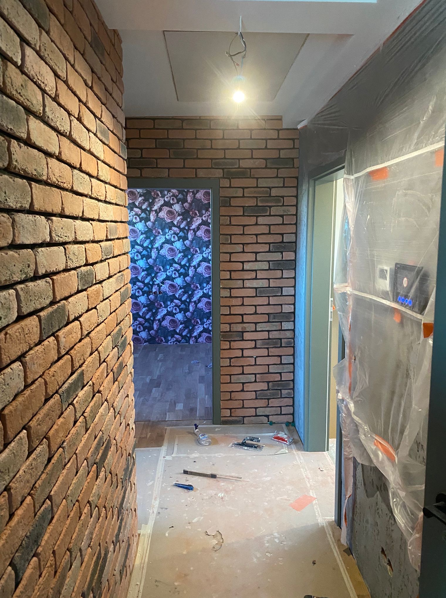 Korytarz po remoncie z ceglaną ścianą i widocznymi narzędziami. Ściana zabezpieczona folią malarską. W tle widoczna ściana z tapetą w kwiaty.