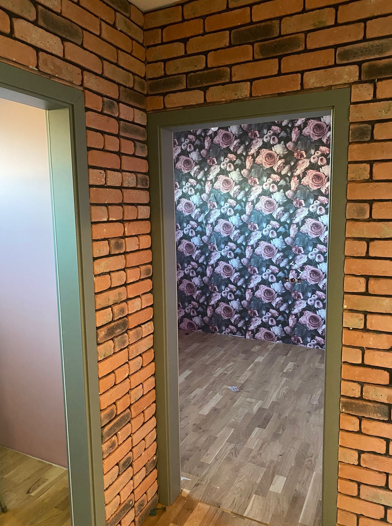 Wykończone wnętrze z ceglaną ścianą i wzorzystą tapetą w kwiaty, widoczne przez futrynę pomalowaną na zielono. Podłoga wyłożona panelami.