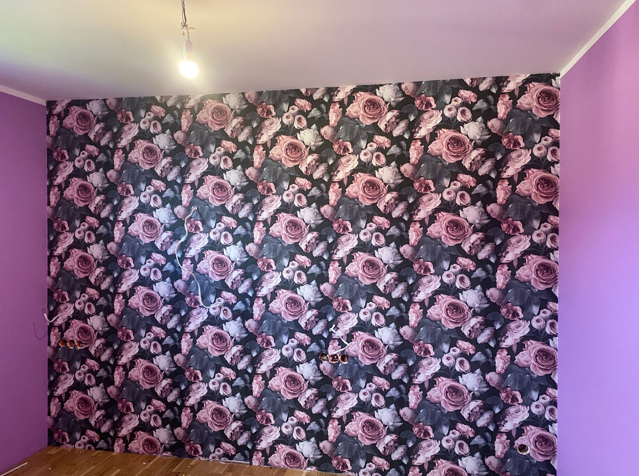 Ściana z tapetą w róże, sąsiadująca z fioletową ścianą. Widoczne wystające kable elektryczne i drewniana podłoga. Oświetlenie sufitowe tymczasowe.