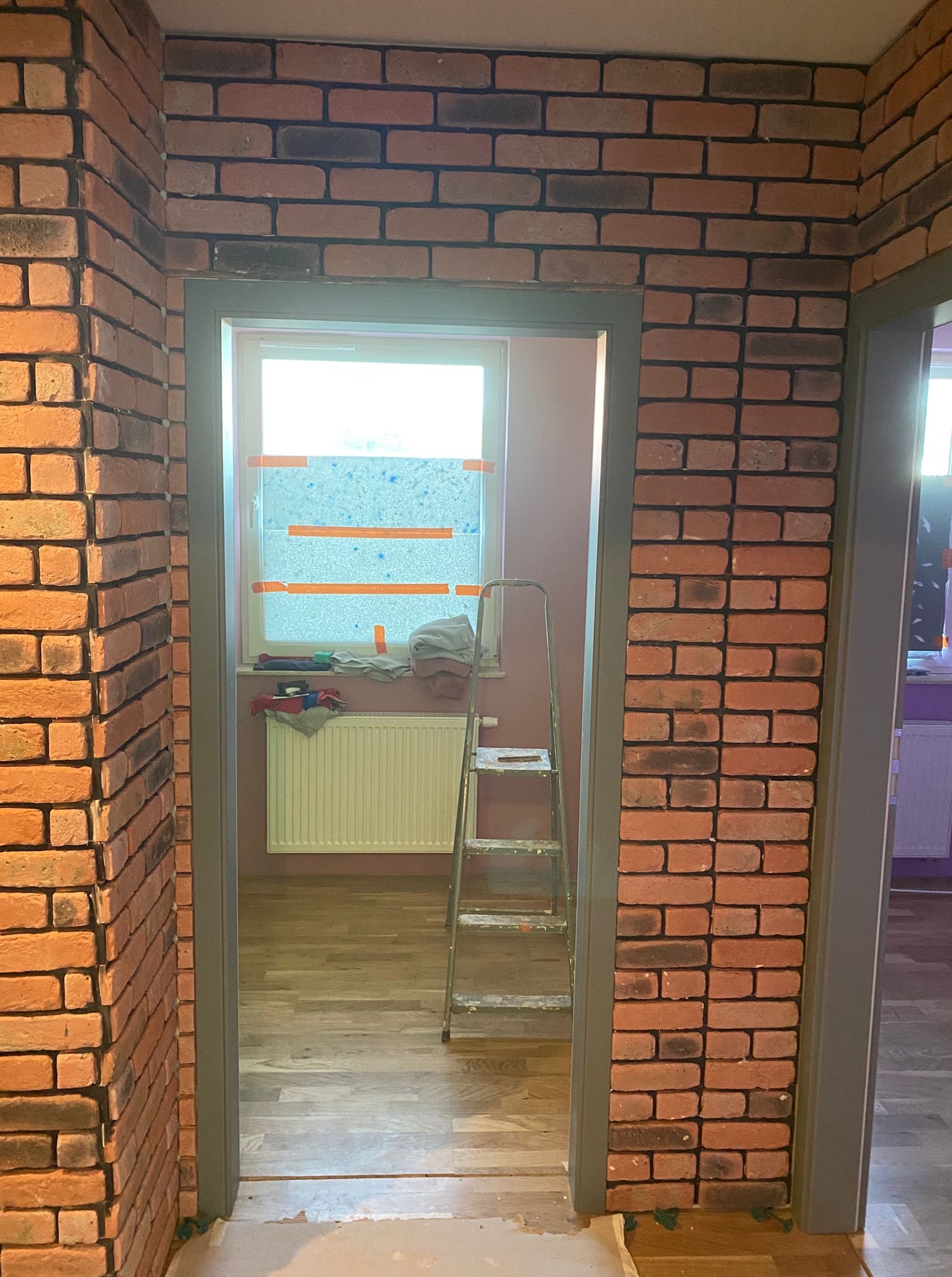 Wnętrze z dekoracyjną ścianą z cegły, widoczne przez futrynę drzwi. Ustawiona drabina malarska, okno z zabezpieczeniem. Podłoga z paneli.