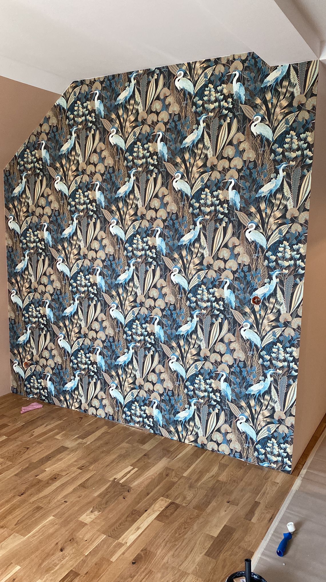 Ściana z wzorzystą tapetą w motywy roślinne i ptaków, świeżo położona na skośnej ścianie poddasza. Widoczna drewniana podłoga i fragment ściany w jednolitym kolorze.