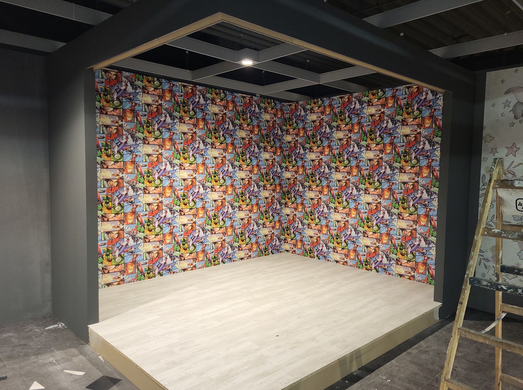 Pokój z podwyższoną podłogą i ścianami w komiksowe wzory. Szare ramy, drabina malarska oparta o ścianę. Nietypowe wykończenie wnętrza.